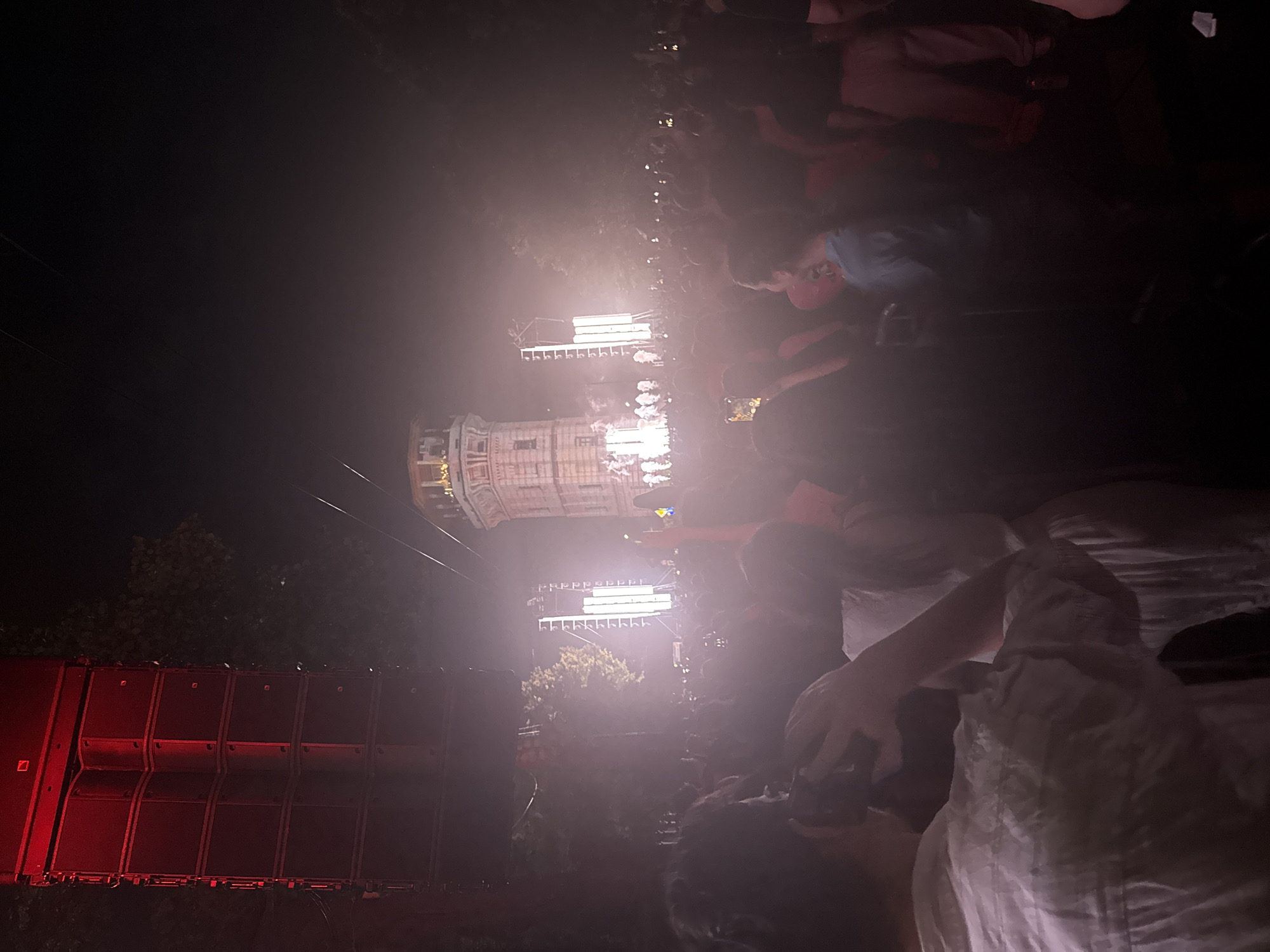 The image depicts an outdoor nighttime concert or event, set against the backdrop of a distinctive, ornate tower structure. The tower, appearing to be made of stone or brick, has multiple levels with windows and architectural details, possibly serving as a landmark or part of a historic building. A large sound system, stacked vertically and illuminated with red lights, is visible to the left of the frame, suggesting a musical performance. Stage lighting is prominent in the center and right of the image, creating a bright, hazy atmosphere. A large crowd of people are gathered in the foreground and mid-ground, many facing the stage and some holding up their phones, presumably to record the event. The individuals are diverse in age and attire, and the overall scene conveys a sense of shared experience and excitement. The presence of trees around the perimeter suggests the event is taking place in a park or open area within a city. The distinctive tower and the general ambiance suggest this scene could be taking place at the Chișinău Water Tower, a historical monument in Chișinău, Moldova, which has been a venue for cultural events.
