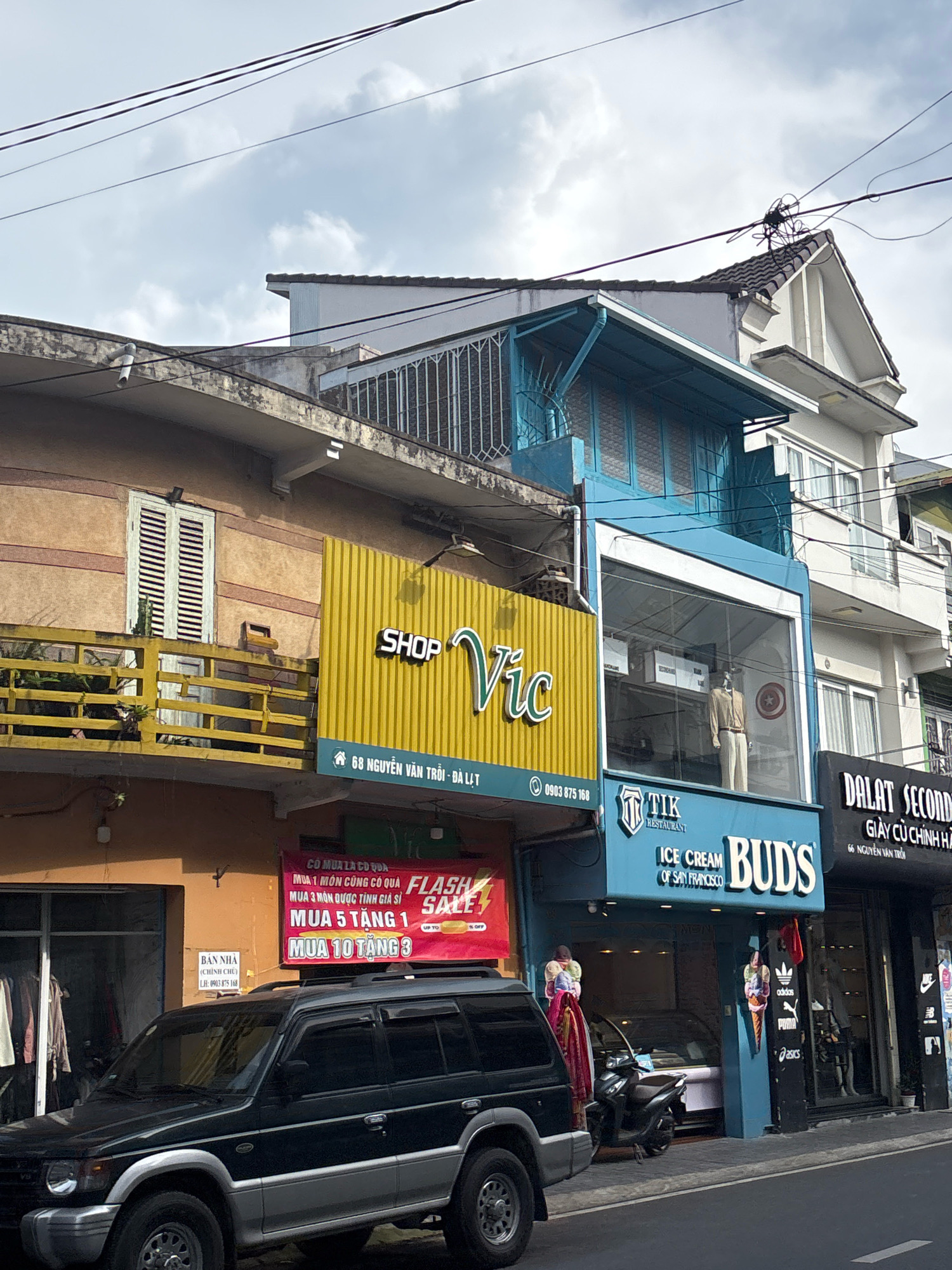 A street scene in Unknown, Unknown shows a row of buildings with shops. In the foreground, a black SUV is parked on the side of the road. The building on the left has a yellow facade and a large yellow sign with the text "SHOP Vic" in white letters. Below this, a red banner with Vietnamese text and "FLASH SALE" is visible. Next to it, a blue building has a large window displaying a mannequin wearing a tan suit. Above the window, a sign reads "TIK RESTAURANT". Further down the street, a building has a sign that says "DALAT SECOND" and "GIÀY CŨ CHÍNH HÃNG". The sky is overcast.