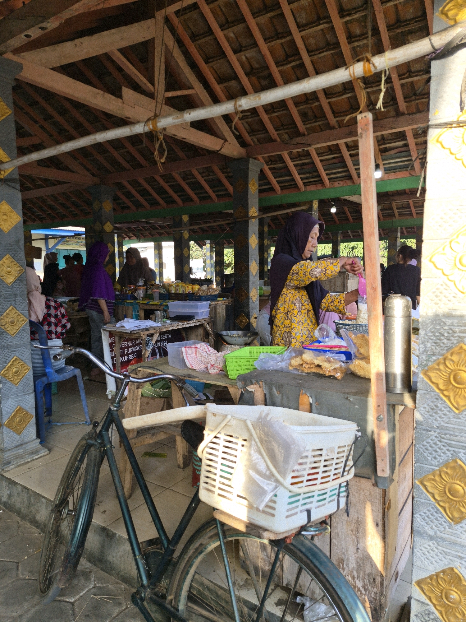 The image depicts an outdoor market stall in Golok, Indonesia, with a woman in a yellow and patterned dress and a dark purple hijab serving customers. She is holding a pink utensil and appears to be scooping something into a plastic bag. The stall is filled with various items, including food, baskets, and containers. A bicycle with a white basket is parked in front of the stall. The market stall is covered by a wooden roof supported by pillars decorated with yellow floral motifs. The ground is paved with interlocking stones. Several other people are visible in the background, some interacting with vendors. The overall scene suggests a bustling local market environment.