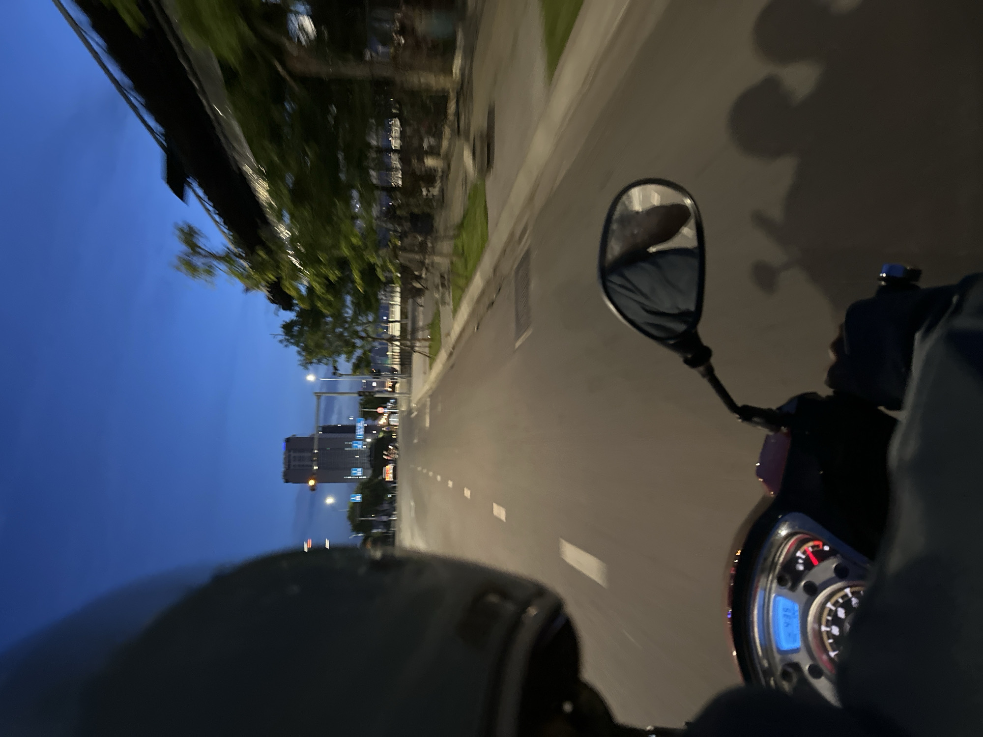The image captures a point-of-view perspective from a motorcycle rider on a street at dusk or night. The rider is wearing a helmet, with the back of it visible in the foreground on the left. The motorcycle's dashboard, displaying the time "24:00", is partially visible on the lower left. The road ahead is paved with lane markings. In the distance, a tall building with illuminated windows stands against a dark blue sky. To the right of the road, there are trees and modern architecture with a reflective, dark-colored roofline. A rearview mirror on the motorcycle reflects the rider's head and torso. Shadows of the rider and possibly another person are cast on the road surface. The scene appears to be in a developed urban area, likely a major road in Đà Nẵng, Vietnam, given the presence of modern buildings and a clear sky often seen in coastal cities.