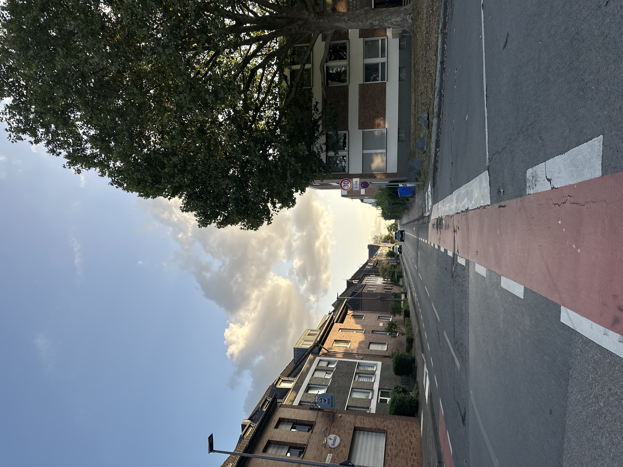 This is a daytime, eye-level, wide-angle outdoor photograph taken on a residential street in Krefeld, Germany, specifically on Bismarckstraße at the intersection with Friedrich-Wilhelm-Straße, near the Krefelder Zoo. The scene depicts a paved road with clear lane markings and a pedestrian crossing marked with red and white paint in the foreground.
On the left side of the street, a row of multi-story brick buildings lines the sidewalk, featuring various window styles and architectural details. Some buildings have signs, including one for "Krombacher" and another that appears to be for a restaurant or business named "Stadt-Schloßbrauerei." A lamppost is visible in front of these buildings.
On the right side of the street, a large, mature deciduous tree with dense green foliage dominates the upper right portion of the frame, casting shadows. Adjacent to the tree is a multi-story building with a brick facade and white horizontal bands. A traffic sign indicating a 30 km/h speed limit and restricted parking hours (7-16h) is posted on a pole near this building. A blue trash bin is also present on the sidewalk.
The road extends into the distance, curving slightly to the left and appearing to ascend. Two cars are visible driving away from the camera on the road. The sky is a light blue with scattered, wispy white clouds, and the lighting suggests late afternoon or early evening sun. The overall environment appears to be a well-maintained urban residential area.