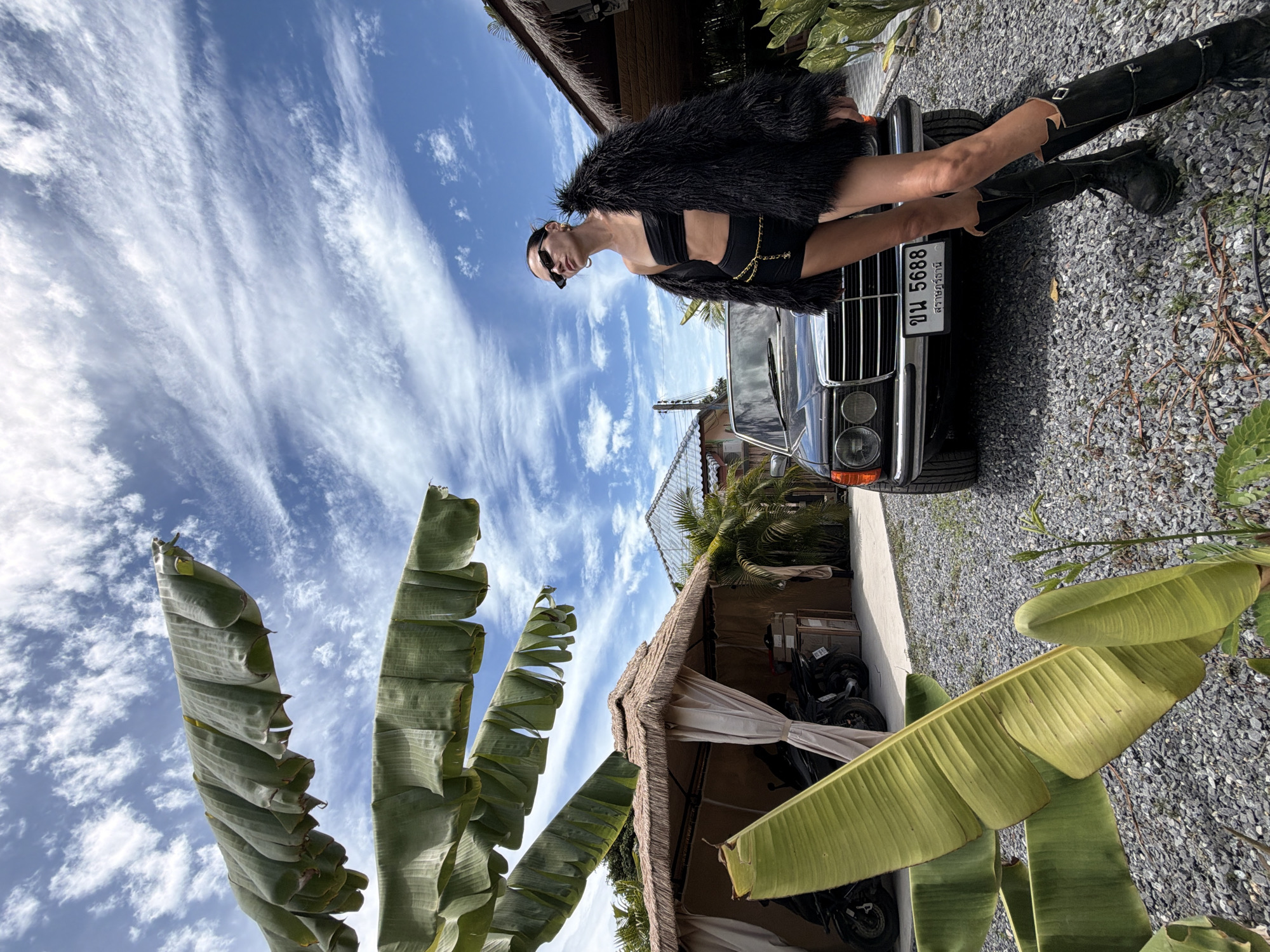 In Ko Samui District, Thailand, a woman stands next to a black vintage Mercedes-Benz sedan on a gravel lot. She is wearing a black bandeau top, black shorts, a black faux fur jacket, and black knee-high boots. Her hair is pulled back, and she is wearing sunglasses and large hoop earrings. The car has a license plate with Thai characters and the number "5688". Behind the car, there are thatched-roof structures with beige curtains, and several motorcycles are parked inside one of them. Large banana leaves hang in the foreground, and the sky above is blue with scattered white clouds.