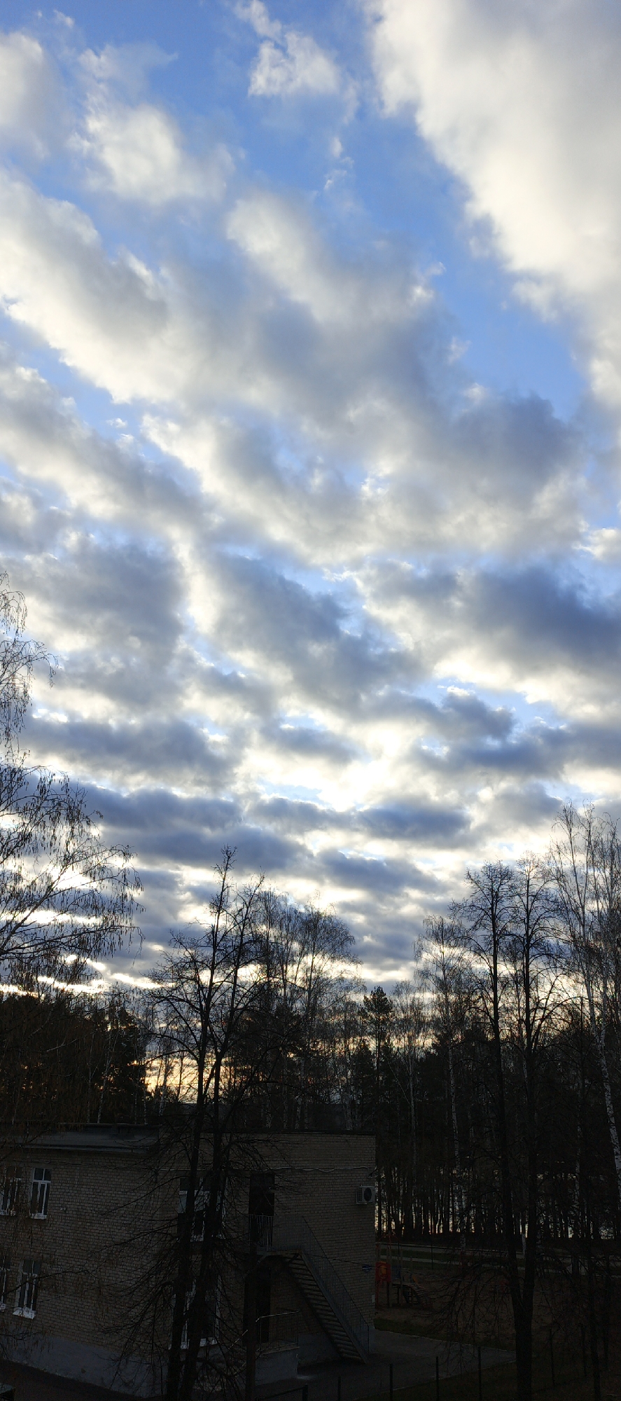 The image shows a cityscape scene in Chaikovsky, Russia. The sky is filled with long, striated clouds exhibiting a mix of white and grey tones with patches of blue visible in between. Below the clouds is a line of trees silhouetted against the sky, indicating a forested area. In the foreground, there is a low, two-story brick building with visible windows and a staircase on the exterior. A portion of a playground is seen behind the building. The overall lighting suggests it could be either dawn or dusk.