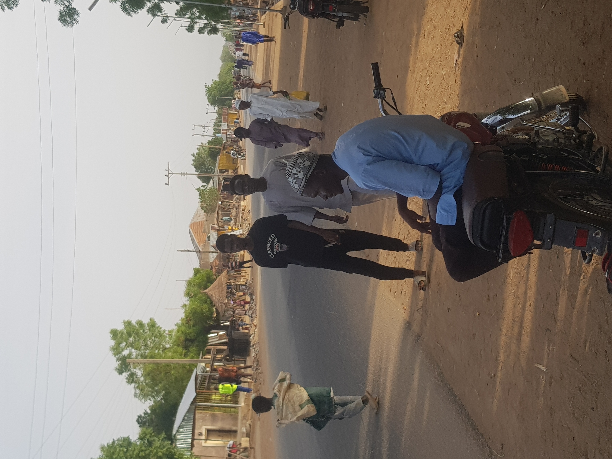 The image shows a group of people gathered around a motorcycle on a street in Dabawa, Nigeria. A man in a light blue shirt is bent over the motorcycle, appearing to be working on it. Two other men stand nearby, observing. One man is wearing a black t-shirt and the other is wearing a white kufi. Other individuals are seen walking along the street in the background, some dressed in traditional clothing. The environment includes buildings, trees, and power lines lining the street. A motorbike is parked in the distance.