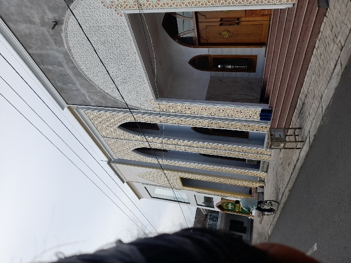 The image shows a mosque located in the city of Kecamatan Tanggulangin, Indonesia. The mosque is a large, modern structure with a gray facade and intricate white designs on the exterior. It has three entrances and a set of stairs leading up to the main entrance. The building appears to be freshly painted. The mosque is located on a street with a paved sidewalk in front. A man is riding a bicycle on the street, and a small trash can is visible on the sidewalk. The sky is clear and blue with a few light clouds. The coordinates of the location are -7.504 latitude and 112.715 longitude. The mosque is likely a place of worship for Muslims in the area.