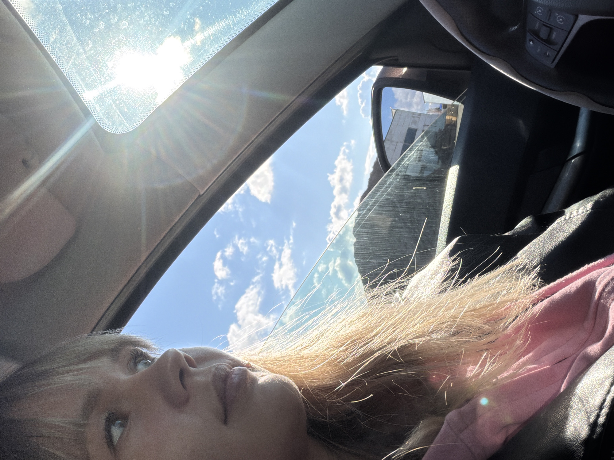 The photograph depicts a woman lying in a car, looking upwards through the sunroof. The sun is visible through the sunroof, with bright lens flare. The sky is blue with white clouds. The woman has blonde hair and is wearing a pink top. The car's interior is visible, including the steering wheel and a side mirror, which reflects a building. The location appears to be urban but is otherwise unknown.