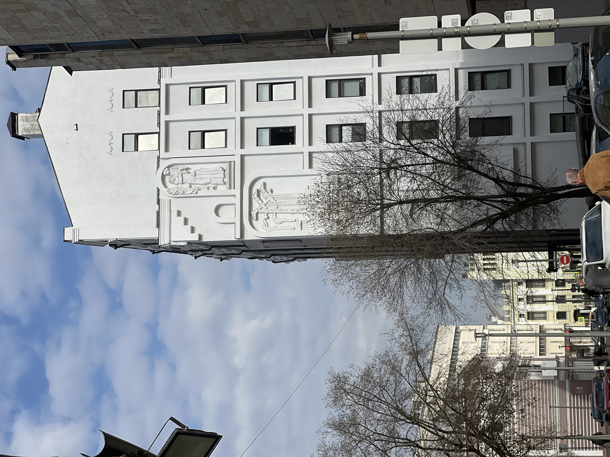 The image shows a view of the Melnikov House in Moscow, Russia, from street level. The iconic white facade of the house dominates the left side of the frame, showcasing its distinctive cylindrical structure and rectangular window grid. Bas-relief carvings adorn the wall. Trees with bare branches are visible in the foreground and partially obscure other buildings on the right side of the frame. A section of blue sky with scattered clouds can be seen above the house and between the buildings. Various street signs are seen and there is a man wearing a brown jacket, and cars on the street.