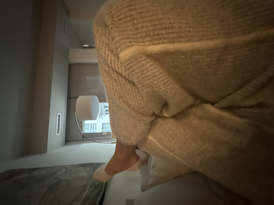 The image depicts an indoor scene, likely a residential unit or hotel room, within an urban environment in the United States; a precise location within Washington cannot be determined. The primary subject is a partially visible person reclining, with their left leg elevated, bent at the knee, and resting on a white pillow. The foot is covered by a white sock. The lower leg and torso are covered by a light beige, textured, knitted blanket or garment. In the background, an abstract artwork with gray and white patterns hangs on the wall. A modern arc floor lamp with a white drum shade stands to the right of the artwork, providing ambient illumination. A window reveals a portion of an exterior multi-story building. A ceiling vent is visible in the upper background. The overall lighting is dim, suggesting evening or low ambient light.