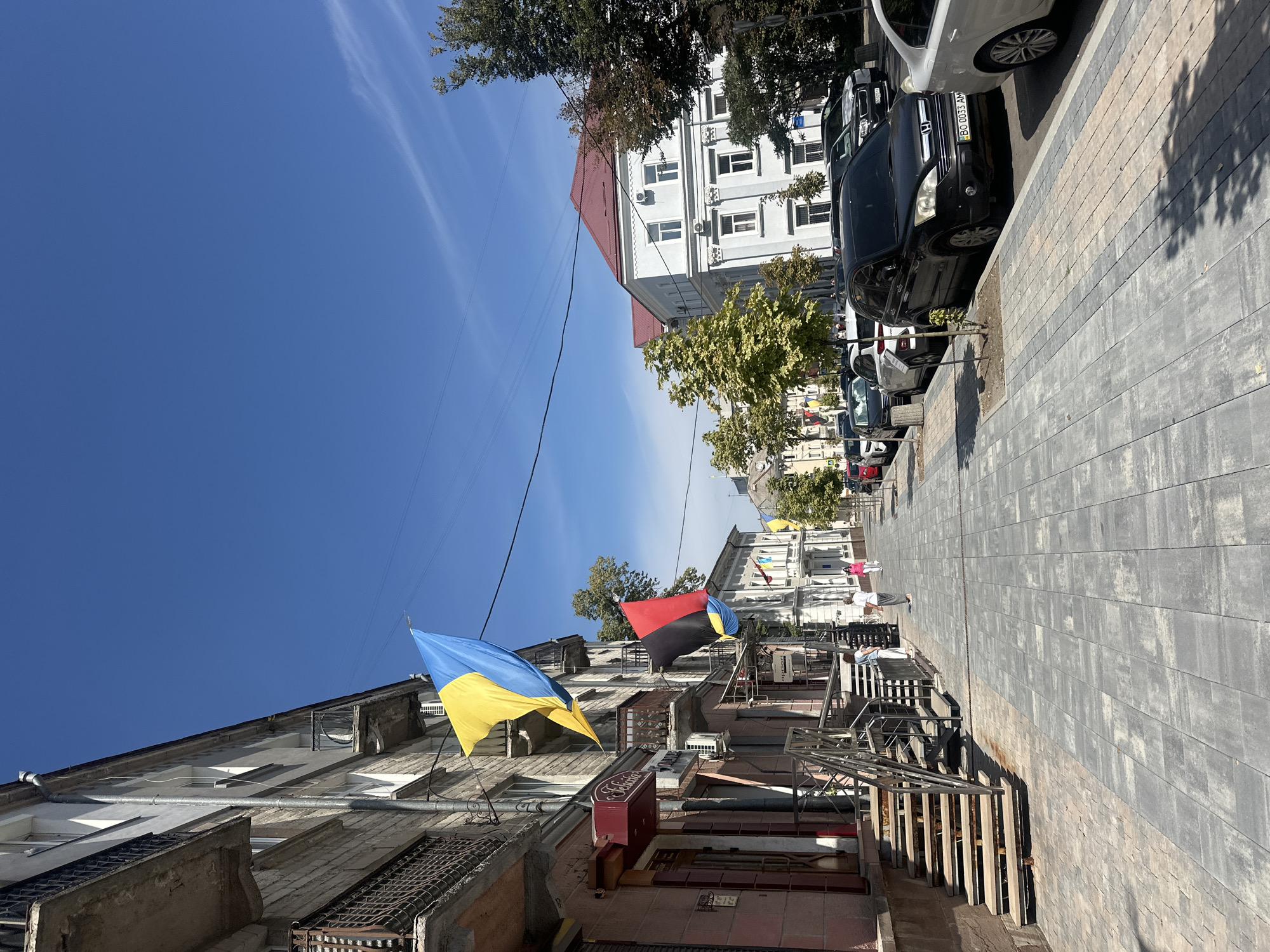 In Ternopil, Ukraine, a sunlit street scene unfolds on a clear day. The perspective is from a sidewalk, looking down a paved street lined with buildings and trees. Ukrainian flags, one blue and yellow, and another black, red, and yellow, are prominently displayed, hanging from the facades of buildings. Several cars are parked along the curb. Two women are walking on the sidewalk in the distance. The architecture appears to be a mix of older and more modern styles. Trees with green and yellowing leaves line the street, and the sky is a vibrant blue with faint wispy clouds. The sidewalk is made of grey rectangular pavers, with some lighter colored pavers interspersed.
