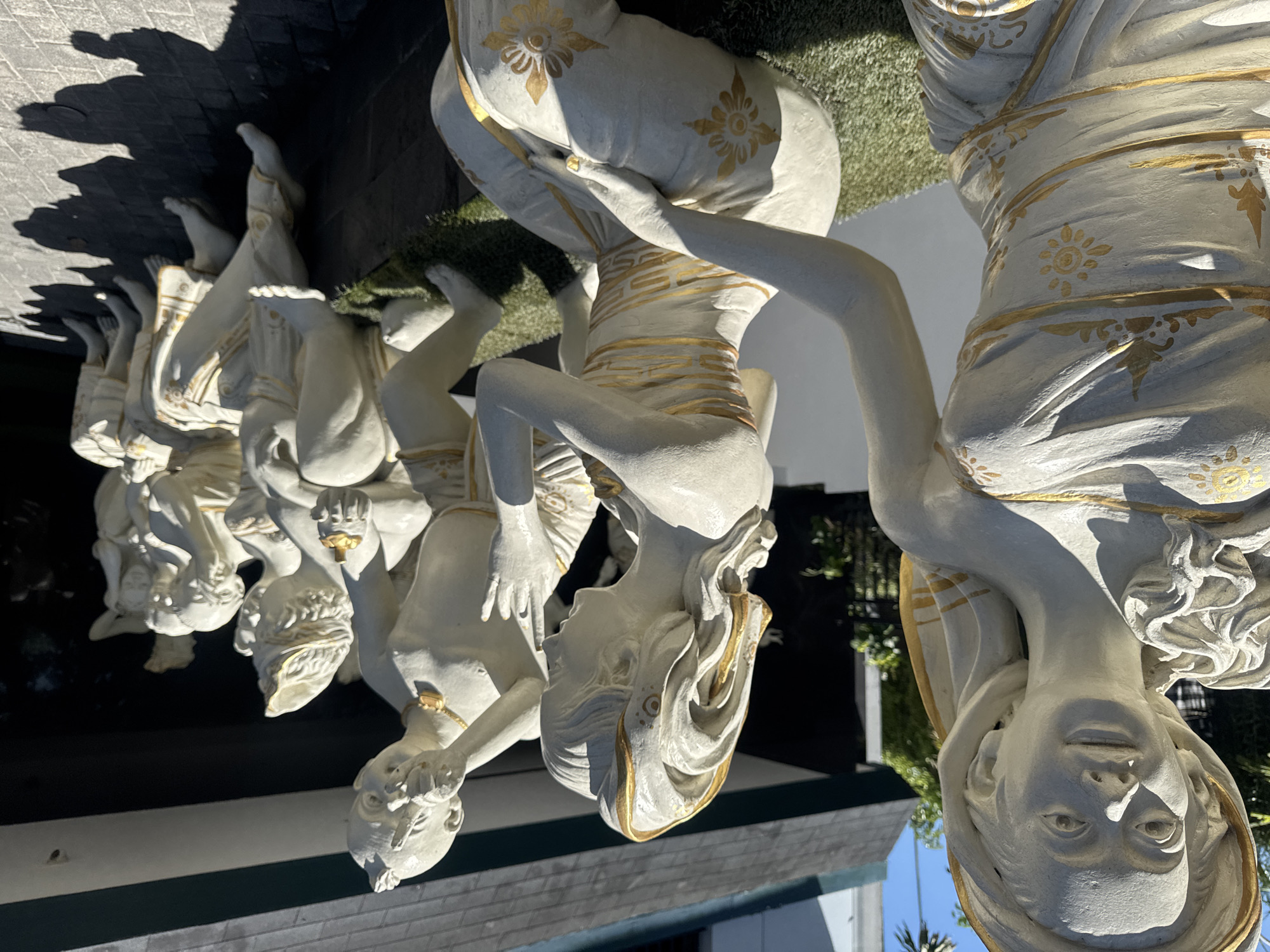 The image shows a row of six white and gold statues depicting kneeling figures supporting a structure. The statues are located in Kuta, Indonesia. The foreground features a close-up view of one of the statues, revealing details like its facial features, drapery, and ornate gold designs. The background shows sections of a building, with trees and clear sky in the distance. The overall lighting suggests a sunny day.