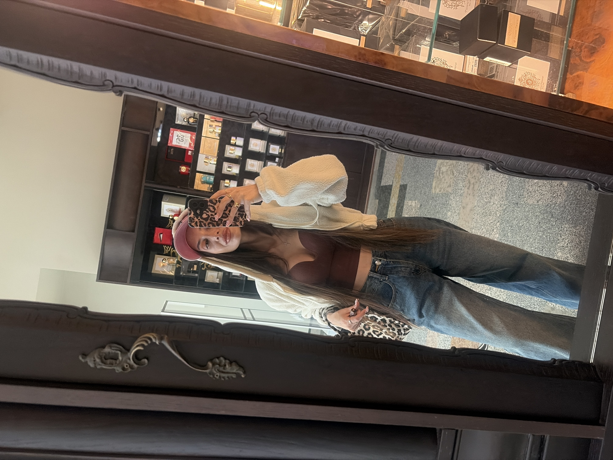 A full-body, medium-shot mirror selfie shows a young woman standing inside what appears to be a shop. The woman is wearing a maroon v-neck crop top, a cream-colored hooded zip-up jacket, light blue wide-leg jeans, and a pink baseball cap. She is holding a phone with a leopard print case in her right hand, taking the photo. A leopard print bag is slung over her left shoulder. She has long, dark hair and is wearing a silver necklace with a cross pendant. The background of the reflection shows shelves stocked with perfume bottles and boxes, suggesting the location is a perfumery or department store. The shop's interior features dark wooden cabinetry and display cases. The woman's reflection is also visible in a large, ornate dark wood-framed mirror, with a dark wooden door with a decorative handle to the left of the frame. The floor is tiled in a grey speckled pattern. The image is likely taken in Saint Petersburg, Russia, given the presence of the perfume brands on display and the overall aesthetic of the shop, which is consistent with high-end retail establishments in the city.