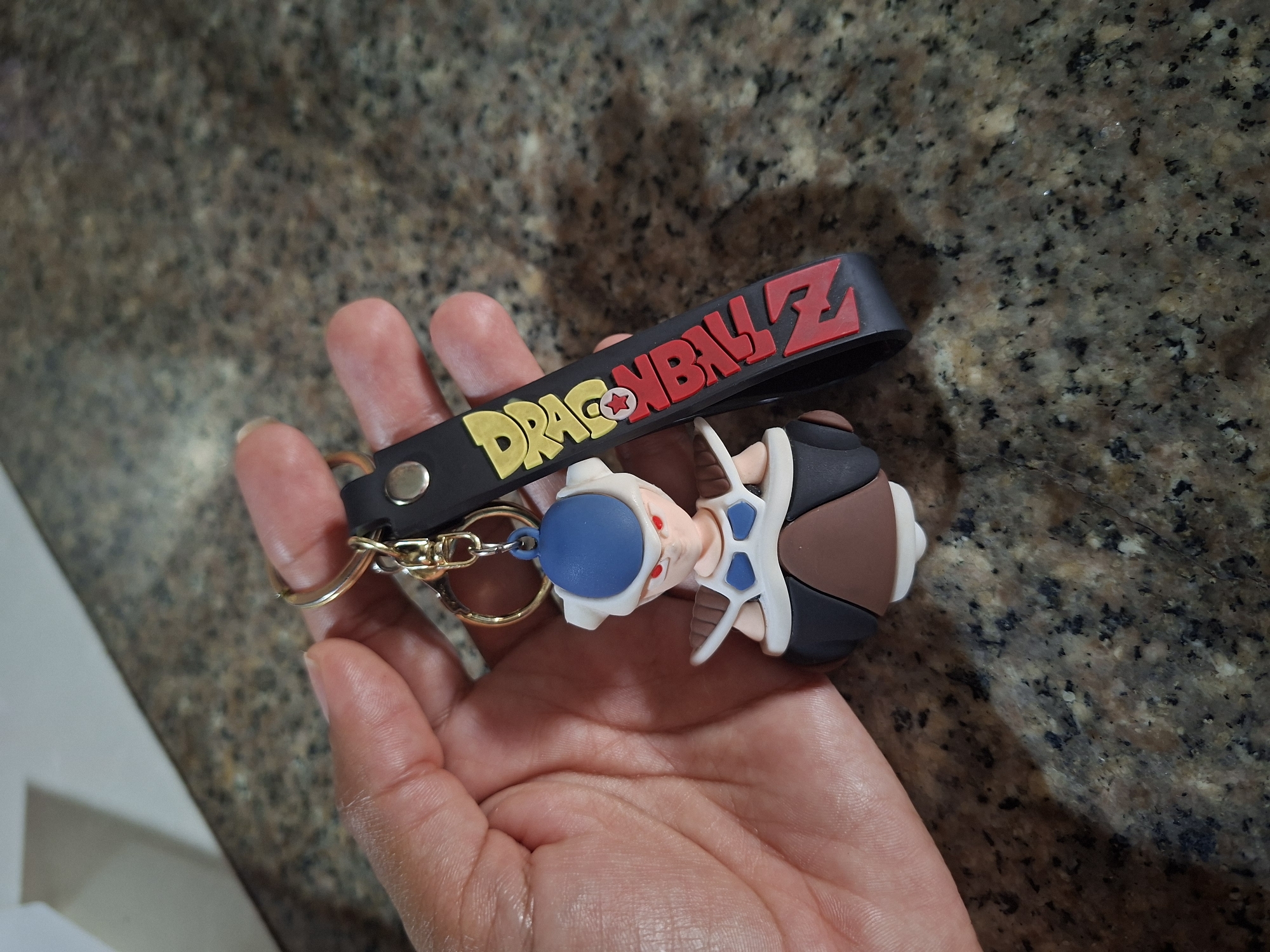A person's hand is holding a keychain with a Dragon Ball Z charm. The charm is of a character with a blue helmet, red eyes, and white and brown armor. The keychain also has a black strap with the words "DRAGON BALL Z" written in yellow and red. The background is a speckled granite countertop. There is no information available to indicate the location of El Valle del Espíritu Santo, Venezuela.