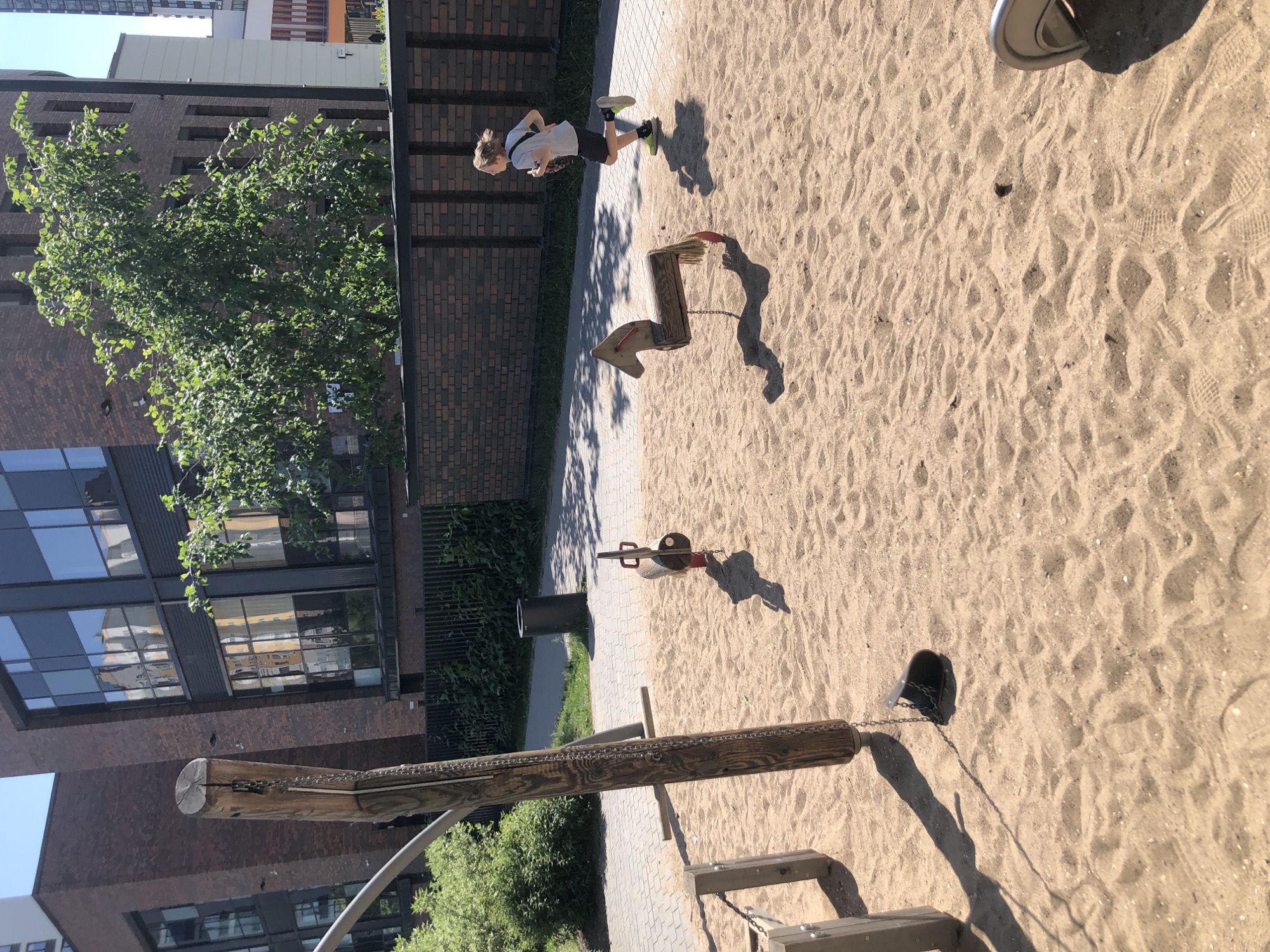 The image shows an outdoor playground with a sand-covered area. In the foreground, a wooden swing set post with a chain hanging down to a detached swing seat on the sand. To the right of the post is a wooden rocking horse toy. In the middle ground, a boy with blonde hair, wearing a white t-shirt, black shorts, and green sneakers, is running from left to right, casting a shadow on the sand. Behind the playground is a paved walkway, a dark metal fence, and a brick wall. Further in the background, there are modern brick buildings with large windows, trees with green foliage, and a tall white building. The sky is clear and blue, suggesting a sunny day. This appears to be a courtyard or residential complex playground. The architecture of the buildings suggests it is likely in Saint Petersburg, Russia.
