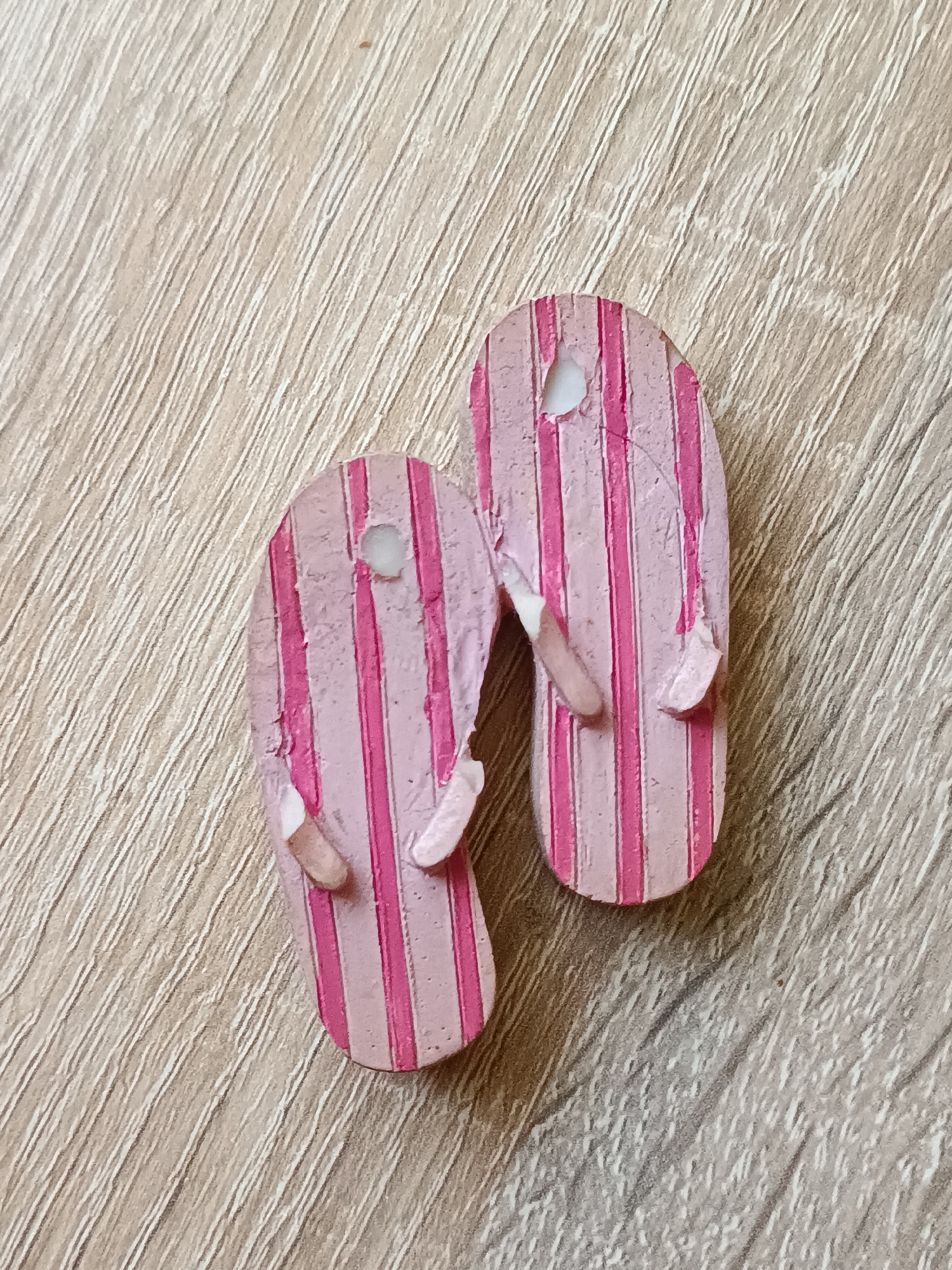 A pair of miniature, pink and white striped flip-flops lie on a wooden surface with a diagonal grain pattern. The flip-flops appear to be made of resin or a similar material and show signs of wear, with small chips and scratches visible. One of the flip-flops has a small hole near the top. The wooden surface is a light brown color with darker lines that create a textured appearance. The scene is lit from above, casting subtle shadows. There are no people or animals present in the image. The location of this scene in Tebras, Latvia, is not determinable from the image itself, as there are no distinguishing landmarks or environmental cues.