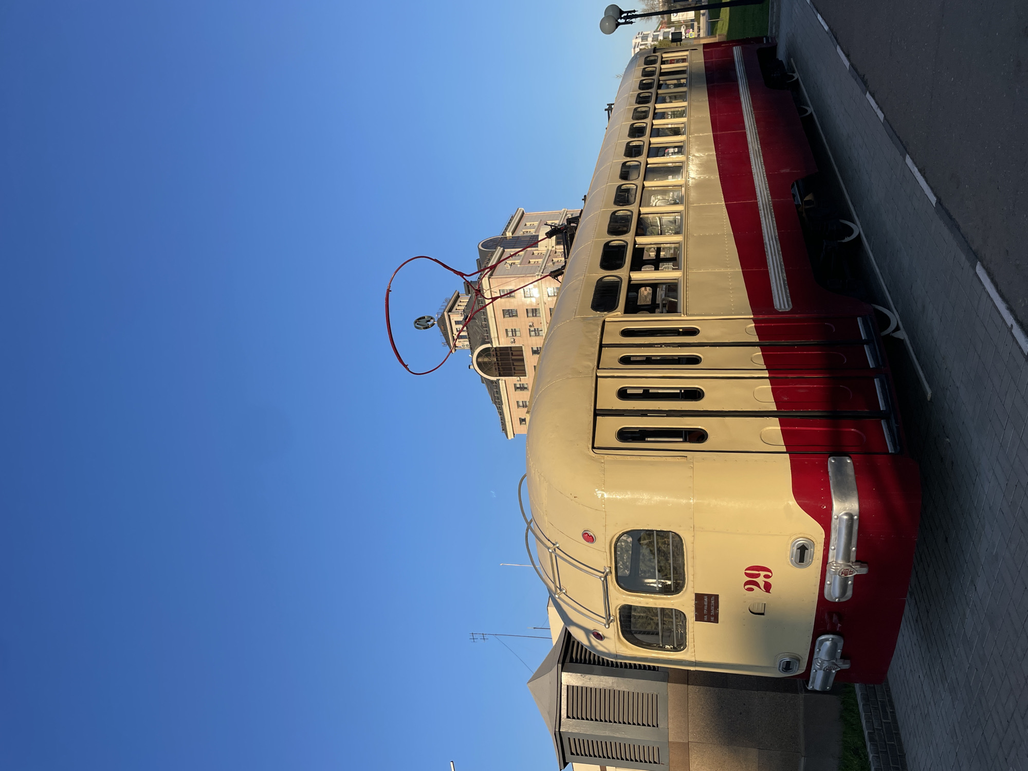 The image shows a vintage trolleybus in Kazan, Russia, with a cream and red exterior. The trolleybus is stationary, parked on a paved surface adjacent to a sidewalk. A building with an arched design stands in the background. The sky is clear and blue.