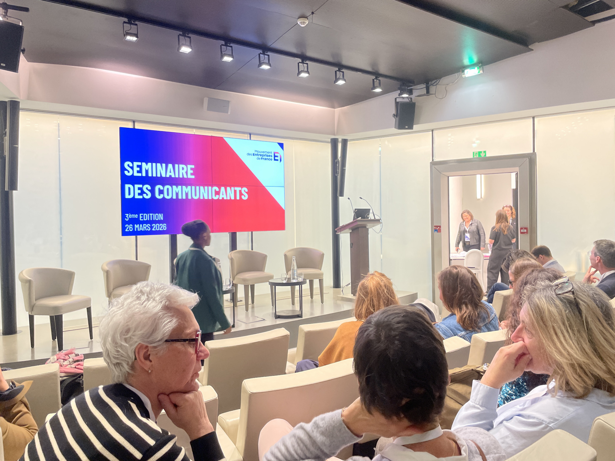 This image depicts an indoor conference or seminar taking place at the Mouvement des Entreprises de France (MEDEF) headquarters in Paris, France. A large digital screen prominently displays "SEMINAIRE DES COMMUNICANTS," "3ème EDITION," and "26 MARS 2026," along with the MEDEF logo.
Approximately a dozen adult attendees are seated in cream-colored chairs, facing the stage and screen. In the immediate foreground, a woman with short white hair and glasses, wearing a black and white striped sweater, rests her chin on her hand, looking right. To her right, a woman with blonde hair also rests her hand on her chin.
On a slightly raised stage area, a person in a dark green jacket walks from left to right. Two cream-colored armchairs are positioned on the stage, flanking a small dark circular table holding a glass water bottle and two empty glasses. To the right of the stage, a wooden podium with microphones and a laptop is visible.
In the background, an open doorway reveals additional individuals, suggesting entry or exit from the room. The venue features a modern design with white walls, glass partitions, and a dark ceiling equipped with track lighting, recessed lights, speakers, a smoke detector, and a green illuminated emergency exit sign.