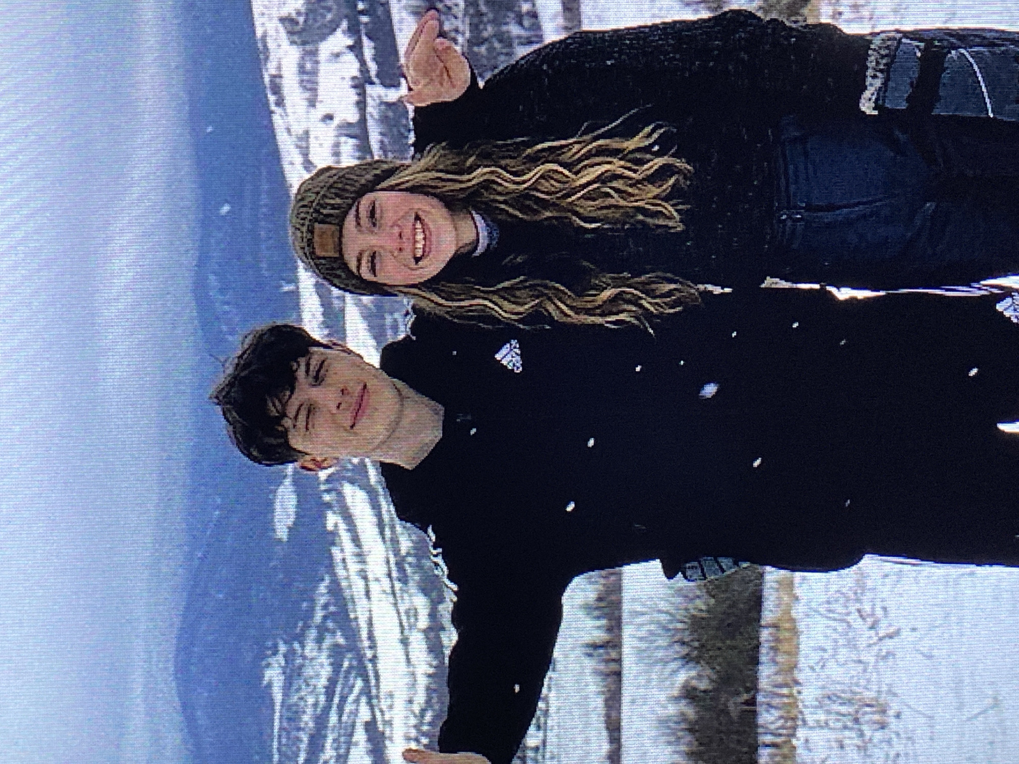 The image shows a young couple standing in a snowy landscape with mountains in the background. The man on the left is wearing a black hooded sweatshirt with white stripes on the sleeves and the Adidas logo. He is smiling and has his arm around the woman on the right. The woman is wearing a dark beanie, a dark sweater, and dark pants. She is also smiling, with her hair down. They are both looking at the camera. The environment is wintery, with snow on the ground and on the mountains. The sky is cloudy and overcast. There is no indication that this location is in Poznan, Poland.