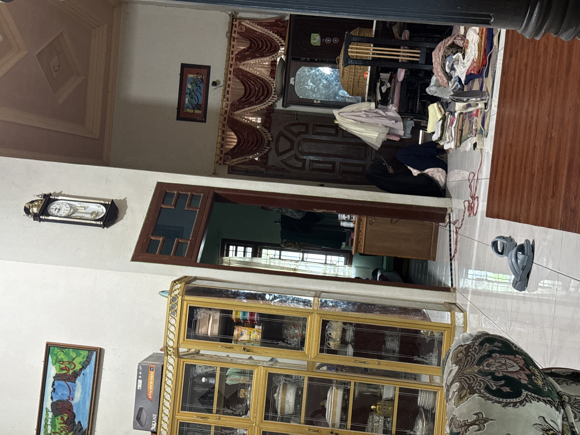 In an indoor setting in Bireuenmeunasahblang, Indonesia, a dimly lit room exhibits domestic clutter. On the left, a tall, ornate golden cabinet with glass doors displays various dishes and decorative items. Adjacent to it, a plush sofa with a patterned fabric is partially visible. The floor is tiled in a light color.
The center of the image features a doorway leading to another room. A large, dark wooden cabinet with a mirror is to the right of this doorway, with several items of clothing draped over a chair in front of it. Next to the cabinet, a stack of folded laundry and an iron are laid out on a rug. A person wearing a black hijab and dark clothing is seated on the floor near the doorway, appearing to be engaged with the laundry.
The walls are painted a neutral color. A framed painting of a waterfall hangs on the wall to the left of the doorway, and another framed piece with Arabic calligraphy is above the red draped curtains on the right. A pendulum clock is mounted on the wall above the doorway. The ceiling is coffered and painted in a soft brown hue. The overall impression is one of a lived-in space with ongoing domestic activities.
