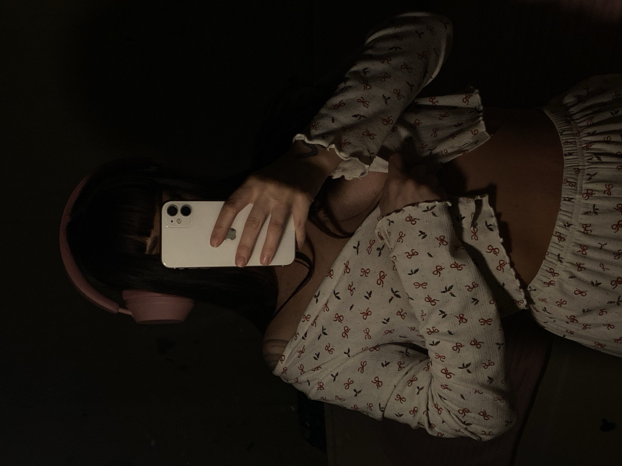 A person is reclining in a low-lit environment, likely indoors. The individual is wearing light-colored pajamas with a repeated pattern of red bows and small birds. They have dark hair and are wearing pink headphones. The person is holding a white smartphone with a visible dual-camera system, positioned as if they are viewing or interacting with the screen. The environment is dark, providing minimal visual information about the surrounding space.
There is no clear indication of the city of Votkinsk, Russia, in this image.