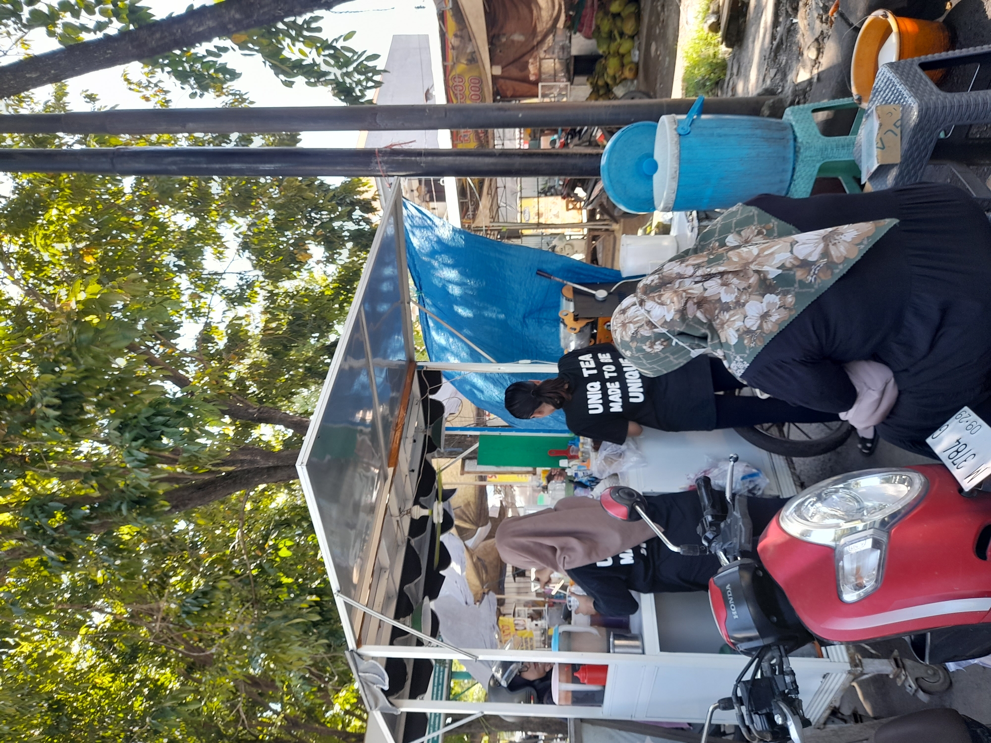 The image depicts a street-side beverage vending operation in Sumokali, Indonesia. Three individuals are working within a mobile cart structure covered by a blue tarp. One person is wearing a hijab with a floral pattern, and another is wearing a black t-shirt with the inscription "UNIQUE TEA MADE TO BE UNIQUE." A red Honda scooter with license plate M 3784 C is parked directly in front of the stand. Blue water containers and plastic seating are visible to the side of the cart. Lush green trees flank the vendor stand, which is next to a street.