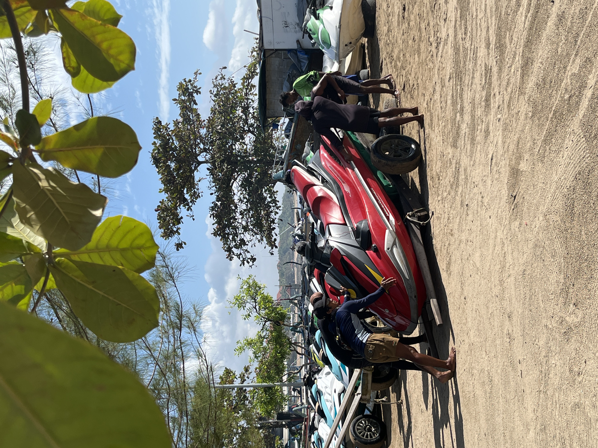 A group of men are gathered around a red jet ski on a sandy beach, likely in Kuta Selatan, Indonesia. The jet ski is positioned on a trailer, and one man in a blue long-sleeved shirt and khaki shorts appears to be pushing it. Another man in a black shirt and shorts is standing next to the jet ski, and a third man in a green shirt and dark shorts is further back. Several other jet skis are visible in the background, lined up on trailers, and a few small structures and trees are also present. The sky is blue with scattered clouds, suggesting it's daytime and possibly a sunny day. The overall scene depicts a recreational area where jet skis are prepared for or have been returned from use.