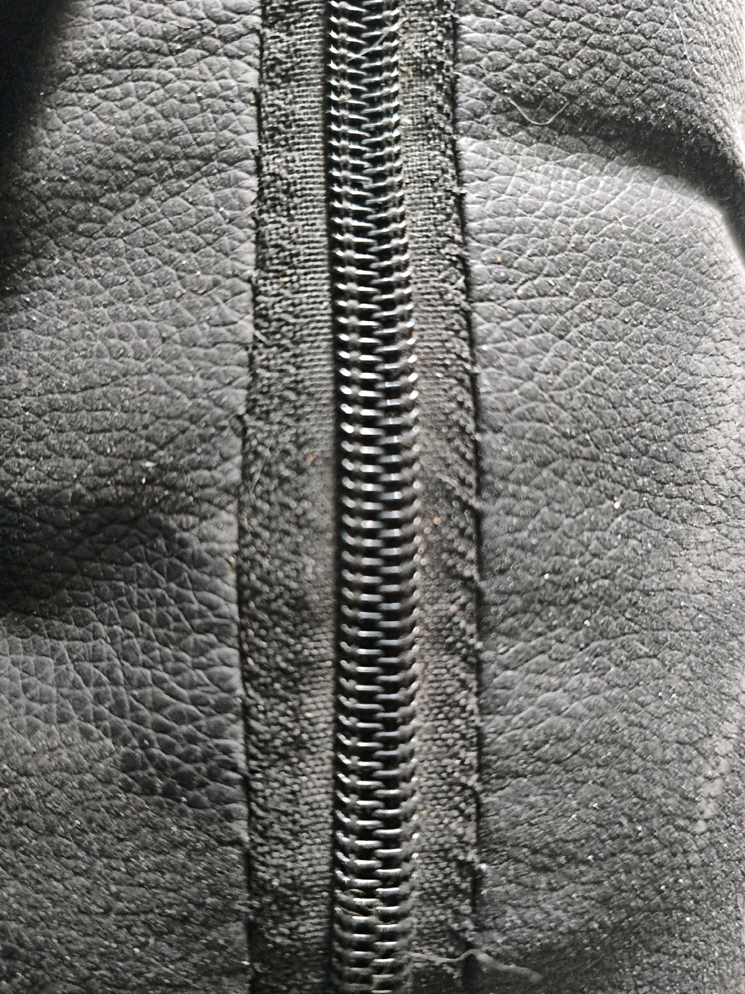 This is a close-up, vertically oriented, high-angle shot of a closed, black zipper on a textured black material, possibly leather or a synthetic imitation. The zipper teeth are metallic and interlocking, forming a continuous line down the center of the frame. The black material on either side of the zipper has a distinct, pebbled texture, with small, raised bumps. The lighting creates shadows that highlight the texture of the material and the depth of the zipper. There are no people, actions, or interactions visible. The image focuses solely on the texture and detail of the zipper and the surrounding material. There is no information in the image to place it in Sukasejati, Indonesia.