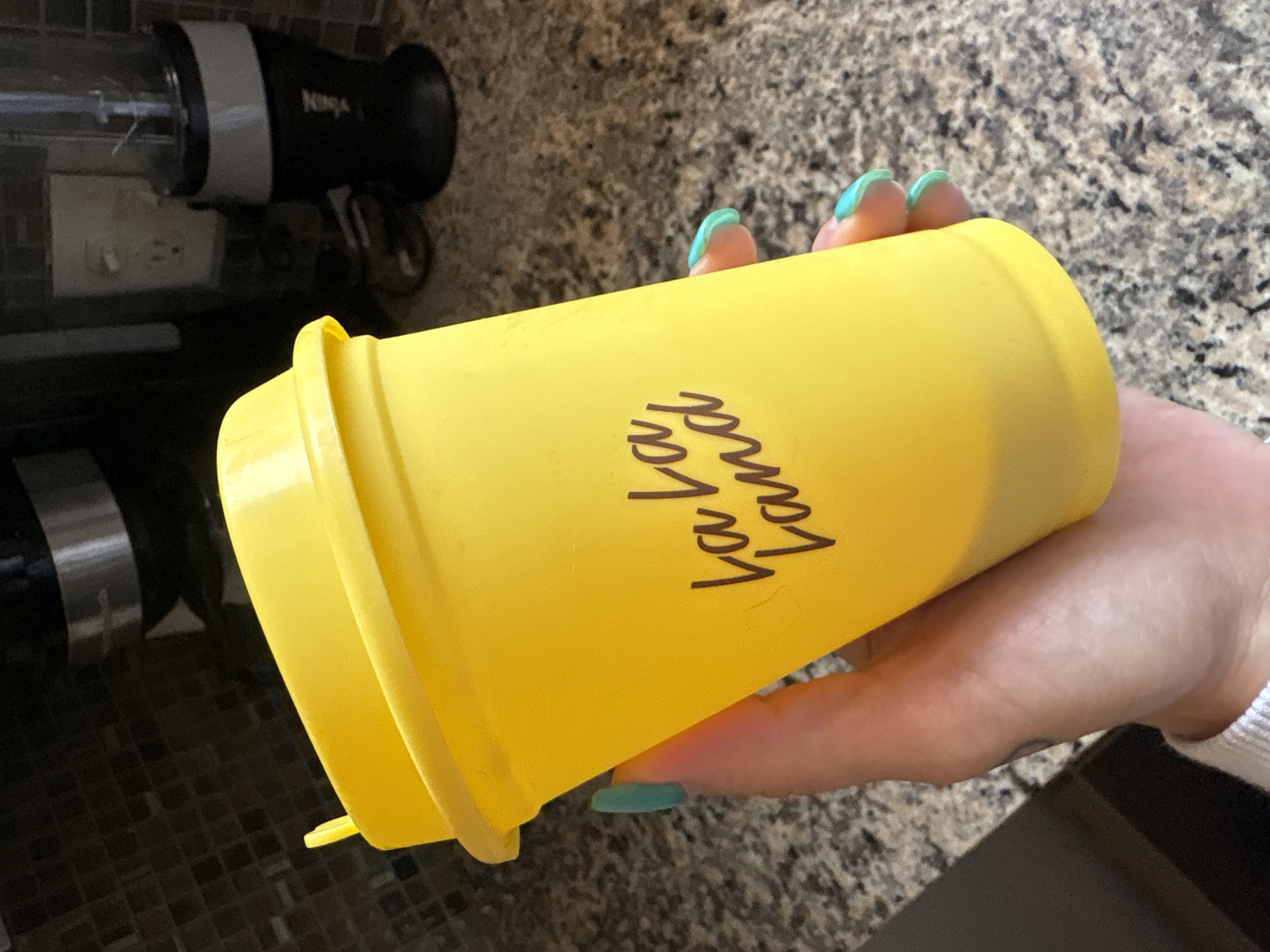 A hand holds a yellow shaker cup with "La La Land" written in dark cursive. The fingernails are painted turquoise. The background shows kitchen appliances, a granite countertop, and tiled backsplash. This image does not allow determination of a location within Seal Beach.