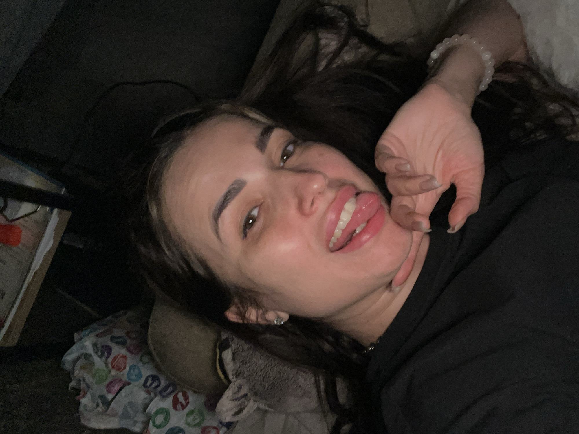 The image shows a woman lying down, presumably indoors in Unknown, Unknown. She is facing the camera with her tongue sticking out. She is wearing a black top and a white beaded bracelet. Her hair is dark and falls around her face and shoulder. Her left hand is near her chin. The background is dark and contains a pillow and bedding.