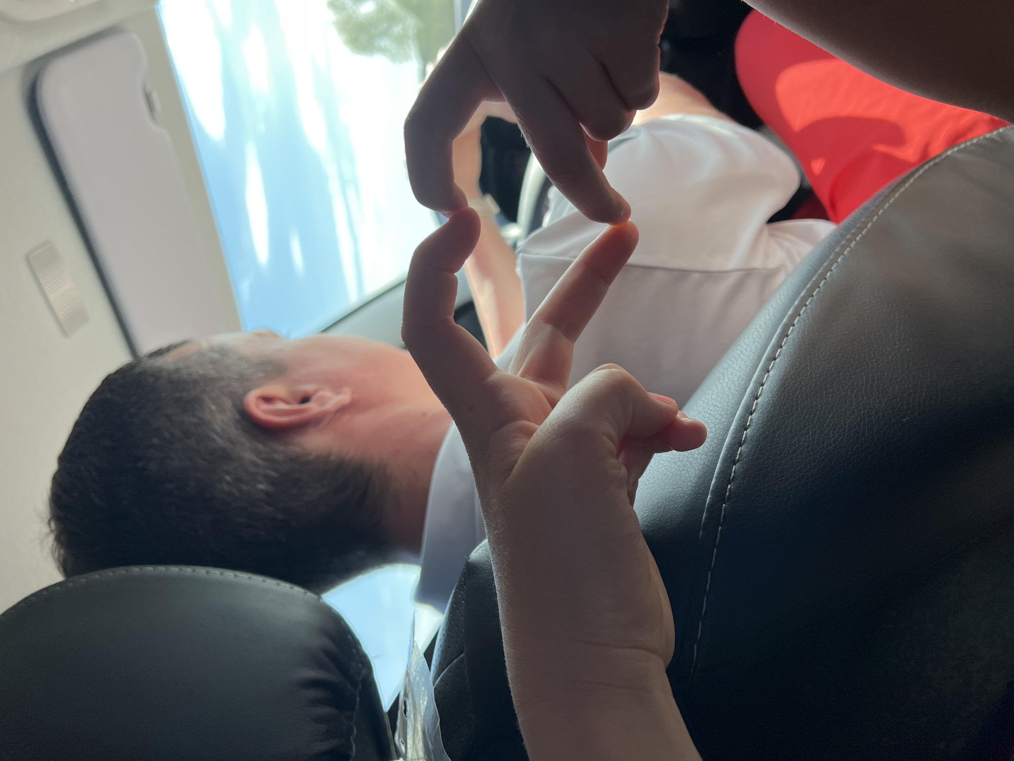 The image depicts the interior of a vehicle, likely a car, with a man reclining in a passenger seat. The background shows a bright, possibly sunlit view through the window, and it is unclear if the location is Niva, Russia. In the foreground, two sets of children's hands are positioned, forming a shape or gesture in front of the man. The man's head is resting against the headrest. The car seats appear to be leather. One of the seated passengers in the rear seat is wearing red clothing.