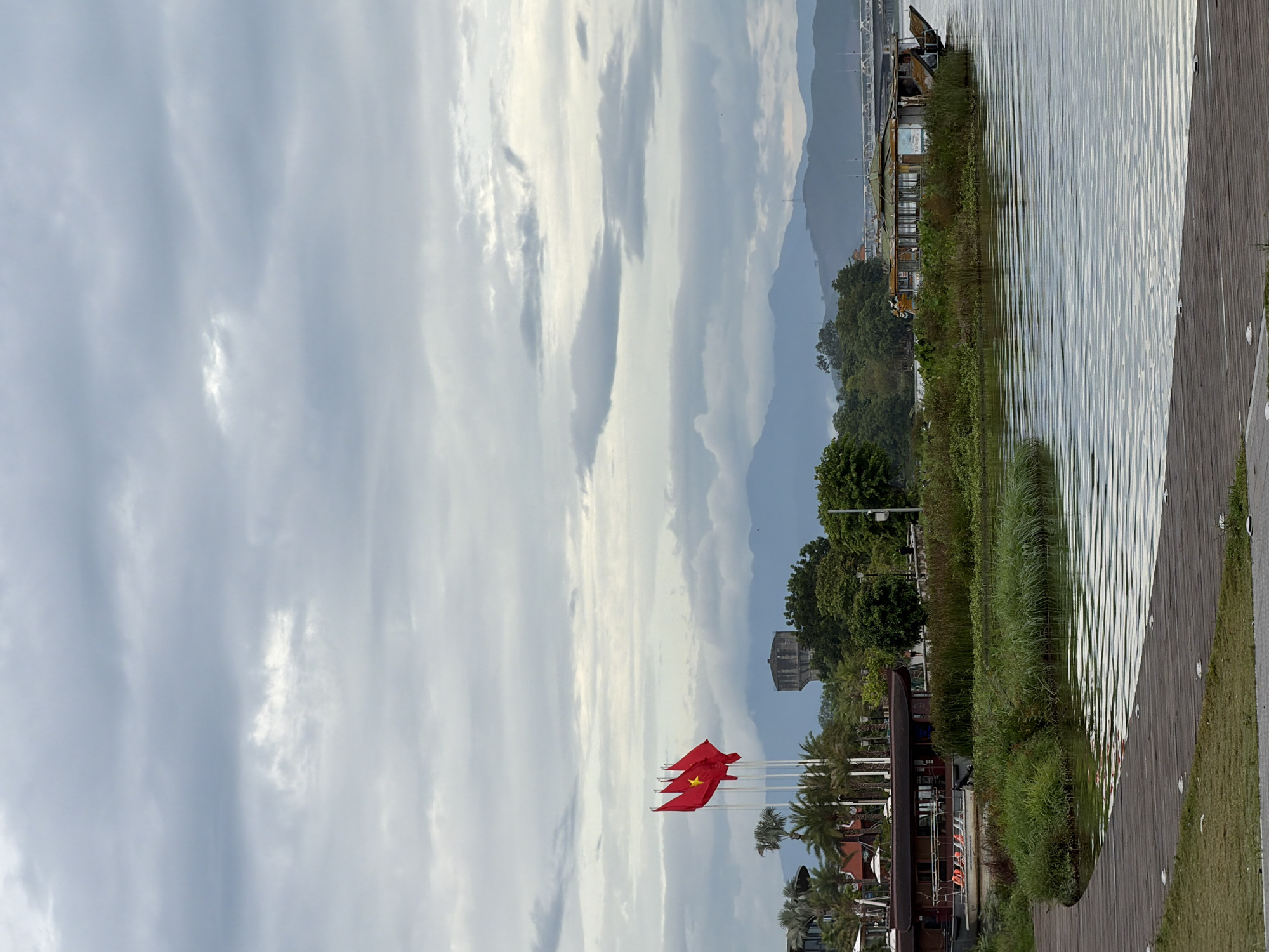 The image captures a waterfront scene in Thuận Hóa Ward, Vietnam. In the foreground, a curved wooden boardwalk is visible, with sections of mowed grass on either side. Beyond the boardwalk lies a body of water with gentle ripples reflecting the overcast sky. On the far bank, dense green vegetation grows along the water's edge.
Several Vietnamese flags, red with a yellow star, fly from tall poles on the left side of the image, positioned in front of a building with white umbrellas and palm trees. A long, brown boat with a covered deck is docked on the right bank, partially obscured by the reeds. In the mid-ground, a large, weathered building, possibly a water tower, stands on a slight incline behind the trees. Further in the distance, a range of blue-hued mountains forms the horizon, topped by a hazy sky with scattered clouds. A bridge, with visible supports and traffic, spans across a waterway in the far right background. The overall atmosphere is muted due to the cloudy weather.