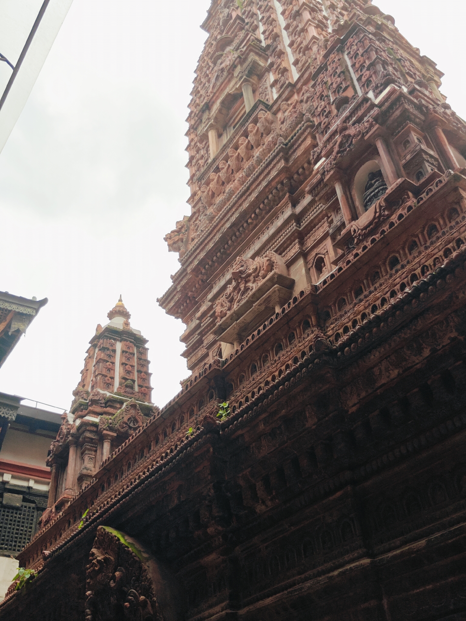 The image shows the exterior of a Hindu temple in Lalitpur, Nepal. The temple is made of red brick and is intricately carved with many decorative elements, including statues and architectural details. The sky is overcast, and there is some greenery growing on the temple walls, suggesting it is outdoors and possibly in a humid environment. The perspective is from a low angle, looking up at the towering structures of the temple. The overall impression is one of grandeur and historical significance, with the detailed craftsmanship of the temple being a prominent feature.