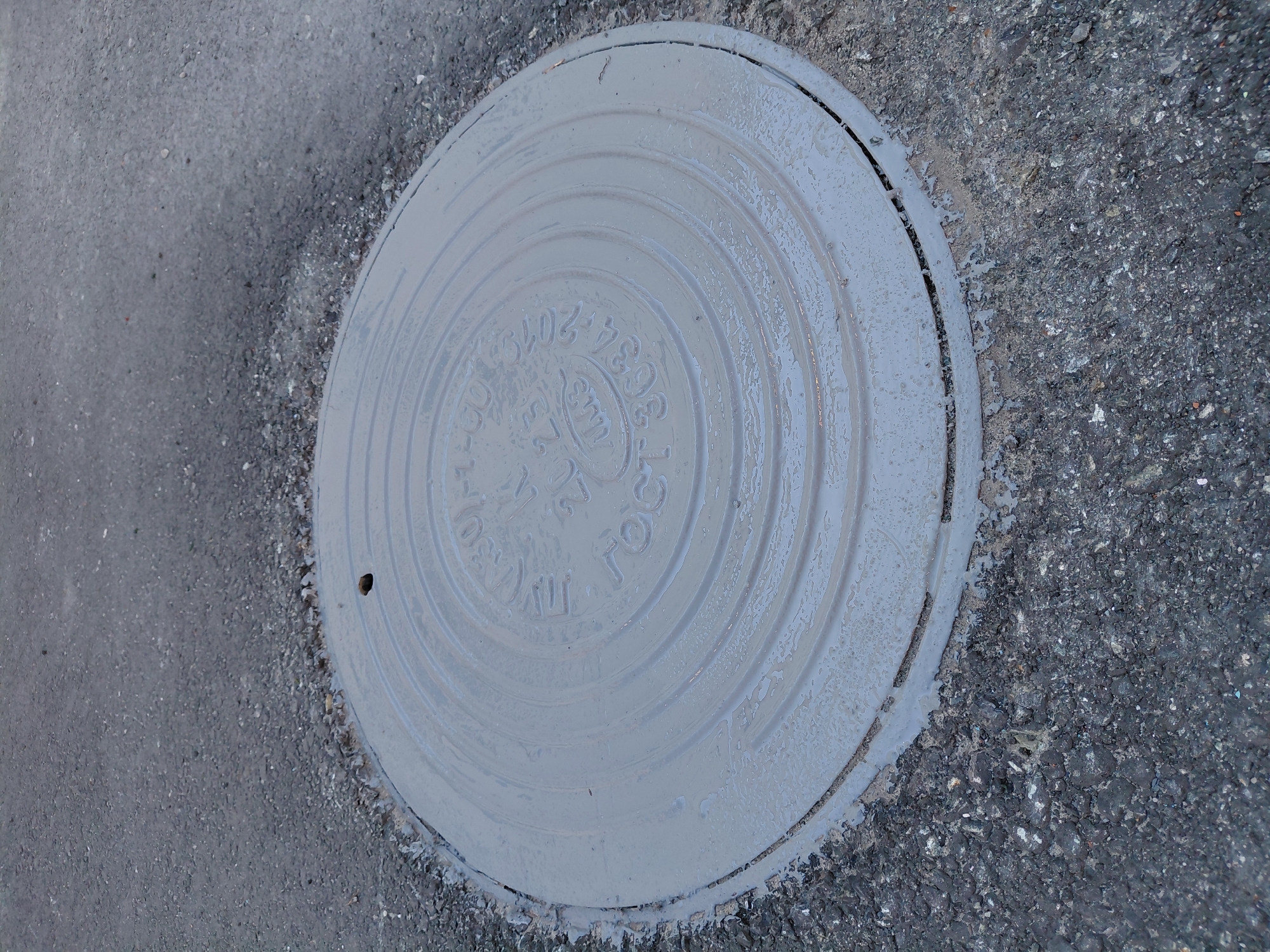 The image displays a close-up, slightly angled view of a light gray, round manhole cover set into dark asphalt pavement. The manhole cover appears recently painted or wet, with some lighter residue visible on its surface and edges where it meets the asphalt.
**Subjects:** The primary subject is a metal manhole cover. The surrounding material is asphalt pavement.
**Objects:** The main object is the manhole cover itself. The textured surface of the asphalt is also prominent.
**Scene, Setting, and Activity:** The scene is an outdoor ground-level utility access point, likely on a street or pathway, given the asphalt. There is no discernible activity taking place; it is a static shot.
**Notable details:**
- **Time of day/Weather:** The lighting suggests natural daylight, possibly overcast or diffused, as there are no strong shadows. The wet appearance of the manhole cover could indicate recent rain or a fresh coat of paint.
- **Location cues:** The text on the manhole cover provides a strong location cue to Russia due to the use of Cyrillic script and the GOST standard.
**Visible text:**
Embossed on the manhole cover, the following text is visible:
- "ГОСТ 3634-2019" (GOST refers to a set of technical standards in Russia and other post-Soviet states, with 3634-2019 likely indicating the specific standard and revision year for manhole covers).
- "ТК" (an abbreviation, possibly for a company or technical specification).
- An emblem or logo, which appears to be a stylized "Им" or "HM" within an oval shape.
- Faint numbers "10-1" are partially visible near the top left.