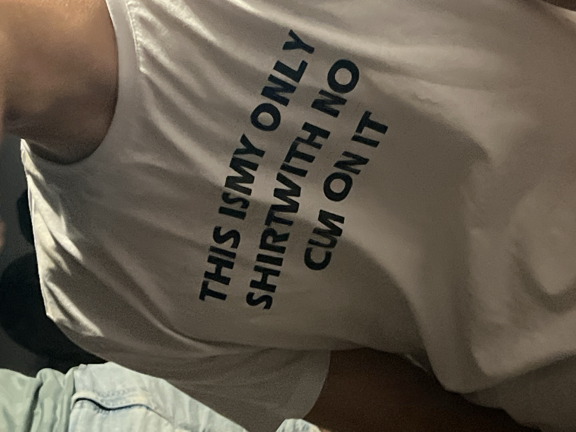 A close-up, medium-shot, low-angle, outdoor, nighttime photograph shows a person wearing a white t-shirt with dark blue lettering. The text on the t-shirt reads "THIS IS MY ONLY SHIRT WITH NO CUM ON IT". The person is partially visible from the neck down, and their arms are not in the frame. The background is dark, suggesting it is nighttime or a dimly lit indoor environment. There are no discernible landmarks or environmental cues to indicate a specific location in Barcelona, Spain, or any other city. The image is focused on the t-shirt and its message.