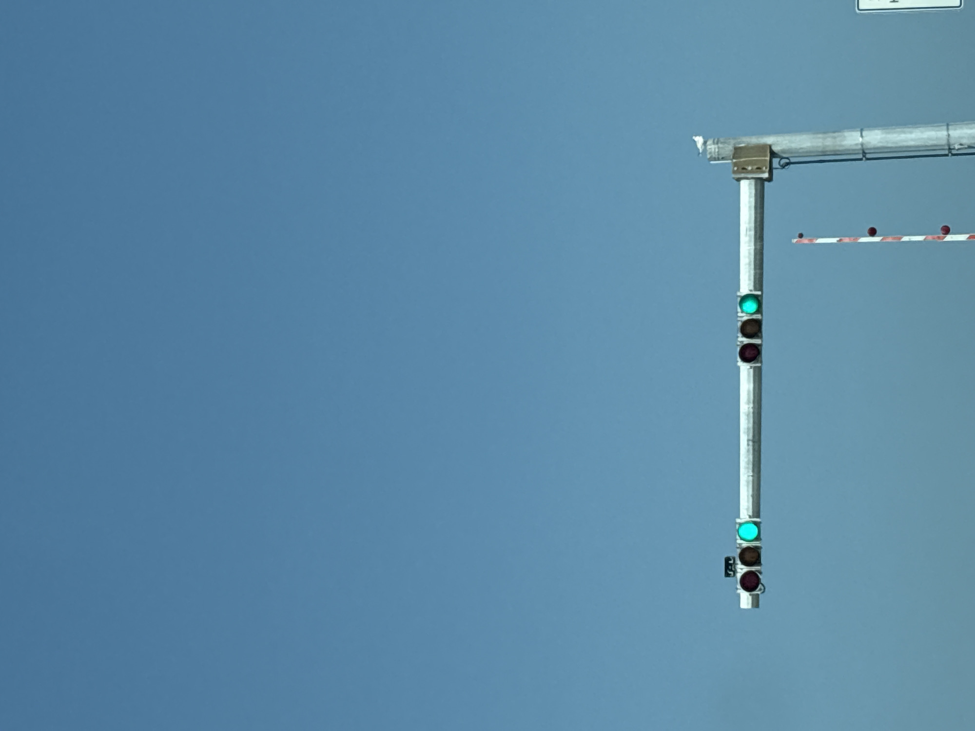 The image shows a traffic signal with a green light illuminated, seen against a clear blue sky. The traffic signal is mounted on a horizontal bar supported by a vertical pole. To the left of the traffic signal, two red lights are visible, and a brown light is between them and the green light. A red and white striped pole with red circular objects is positioned below and to the right of the traffic signal. A white bird is perched on top of the vertical pole. A white sign with black lettering is partially visible on the far right of the image. The scene appears to be a traffic intersection or a railway crossing in Bradenton, Florida, United States of America.