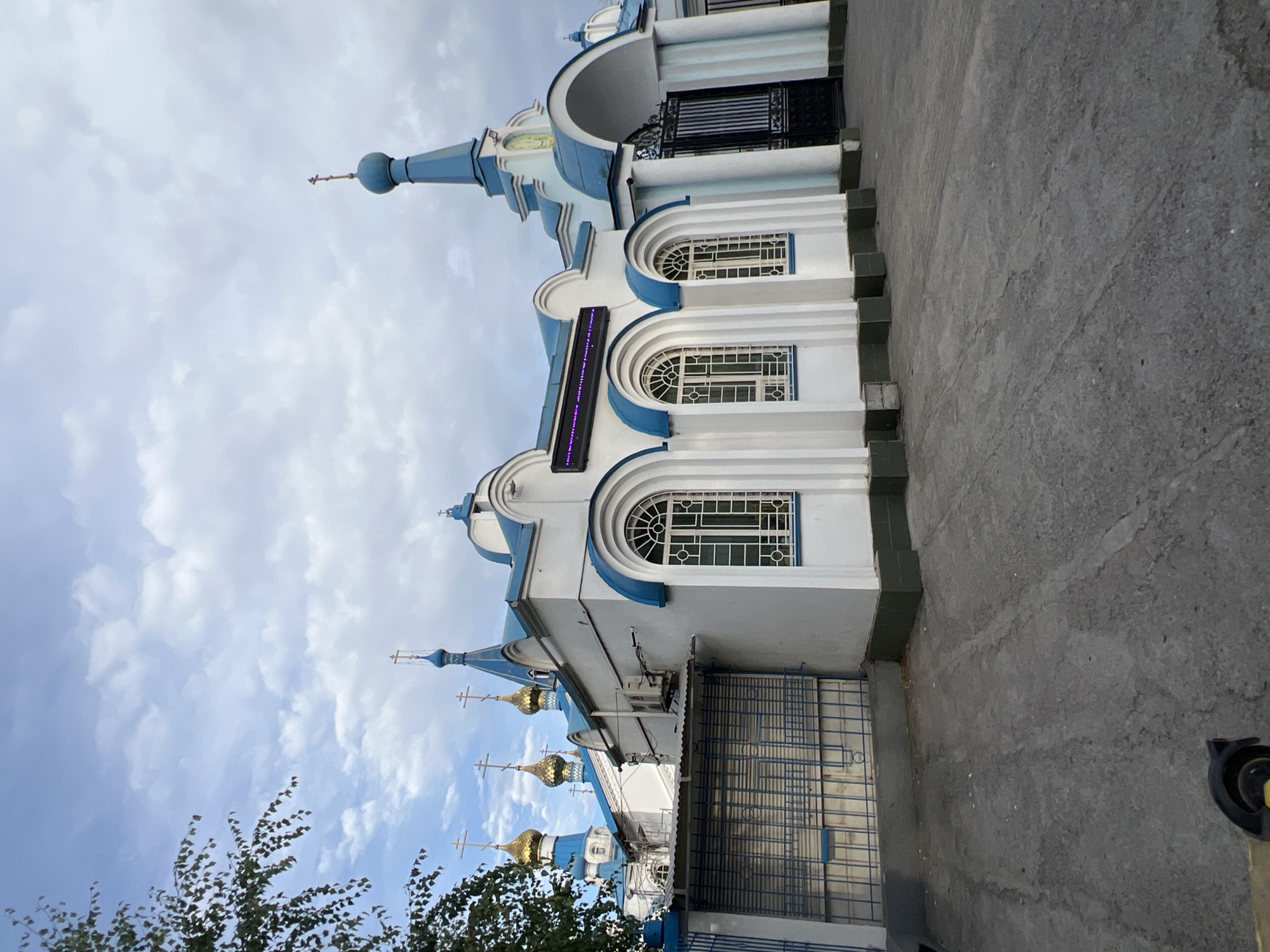 A bright, white and blue Orthodox church stands under a cloudy sky in Bishkek, Kyrgyzstan. The church has multiple golden domes and a tall blue spire. A large black metal gate is visible to the right of the church, and a fence is to the left. The ground in front of the church is paved with asphalt. A tree with green leaves is seen in the upper left corner of the image.
