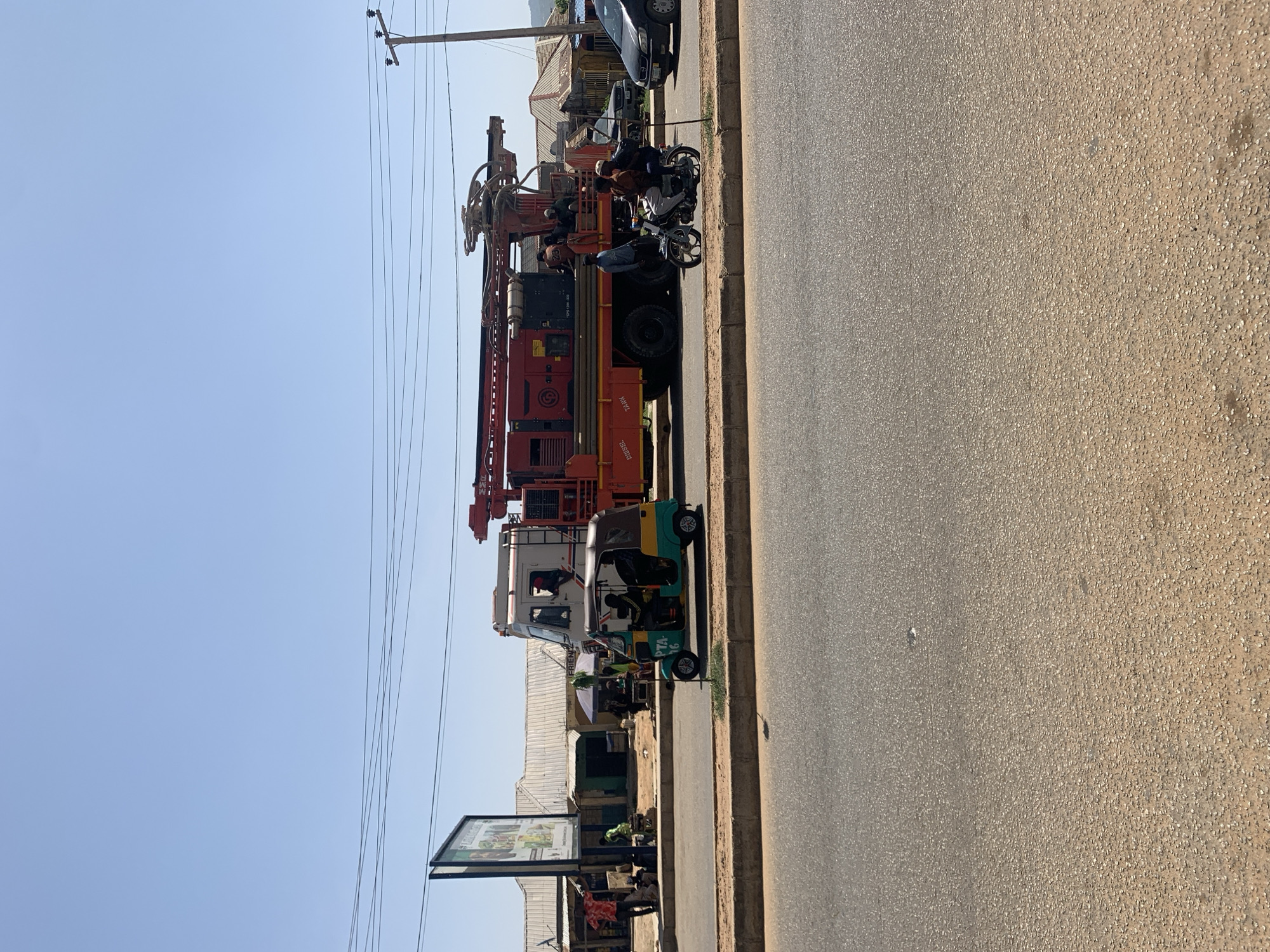 The image depicts a street scene in Rafin Par, Nigeria. A large drilling rig truck is parked on the side of the road. A motorcycle with multiple riders is stopped near the truck. A three-wheeled vehicle (tuk-tuk) is positioned further down the road. Adjacent to the road, there is a sidewalk with shops and a billboard. The sky is clear.