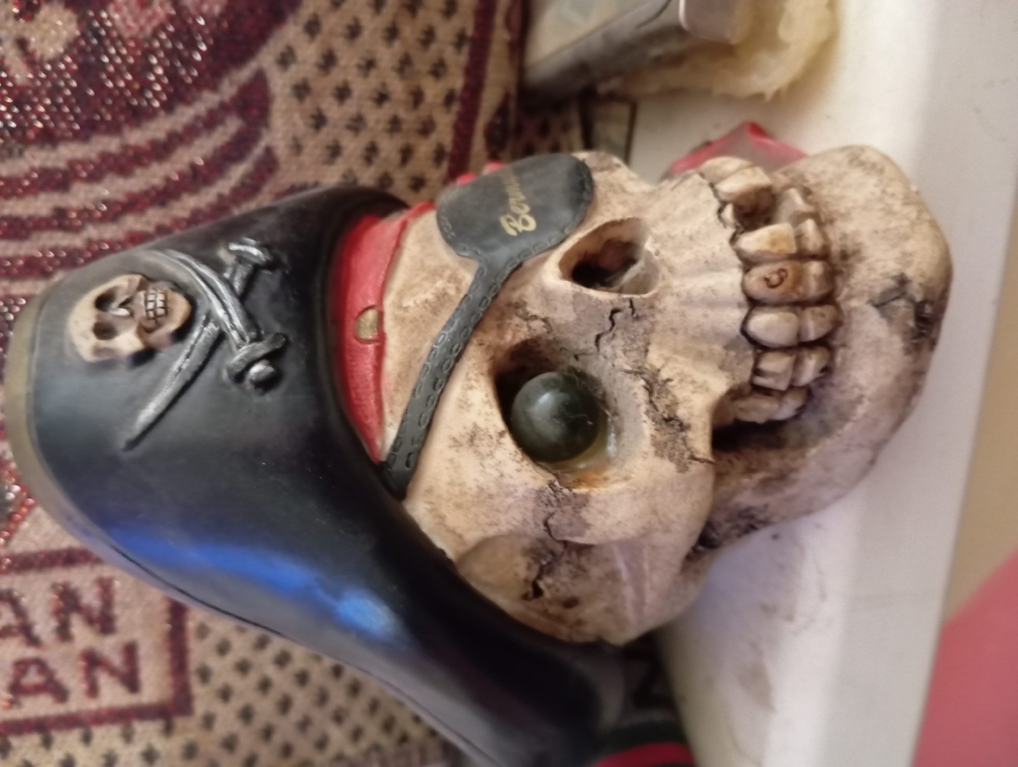 A close-up, eye-level, medium shot captures a resin sculpture of a skull wearing a black pirate hat adorned with a skull and crossbones emblem. The skull has a single green glass eye, a black leather eye patch inscribed with "Bermuda" in gold lettering, and visible teeth. The background consists of a patterned textile. The scene is set on a white shelf. The exact location in Aktau, Kazakhstan, is not determinable from the image.