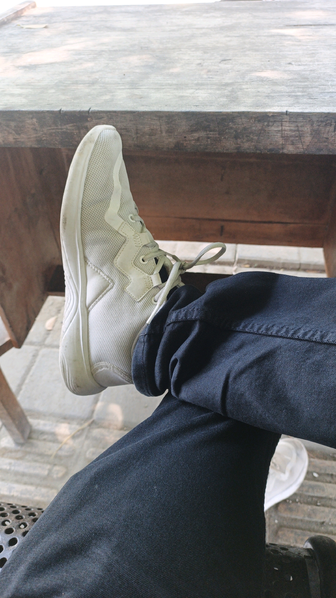 A first-person perspective shot captures a person's legs, clad in dark blue pants, crossed with one foot resting on a wooden table. The foot is clad in a white, mesh athletic shoe with light gray laces. The wooden table appears weathered and aged, with visible grain and occasional dark stains, suggesting it might be an outdoor dining or seating area. The background reveals a tiled floor and the base of a dark metal object, possibly a chair or table leg, hinting at a casual outdoor setting. While the specific location within Denpasar, Indonesia, cannot be definitively identified from the image alone, the overall ambiance suggests a relaxed, perhaps cafe or park-like environment common in urban areas of the city.