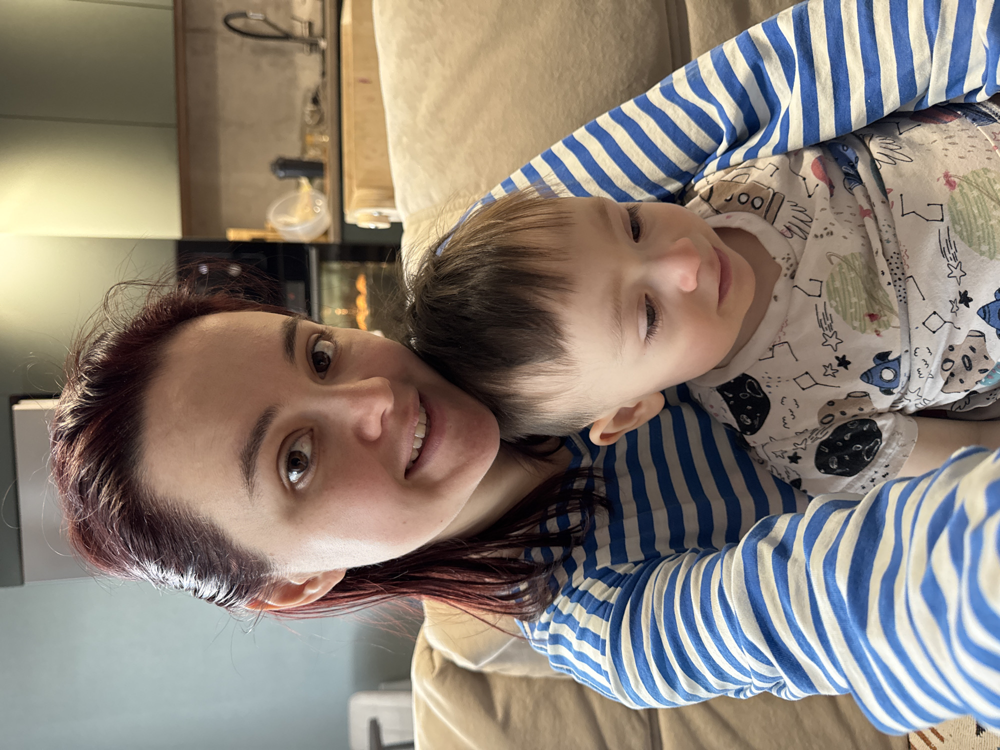 An indoor selfie-style portrait captures a woman and a young child sharing an embrace. The image is oriented vertically, showing the subjects from the chest up.
The woman, with reddish-brown hair, is on the left, looking directly at the camera with a warm smile. She has dark eyebrows and brown eyes. Her face shows a pleasant, content expression. She is wearing a long-sleeved shirt with horizontal blue and white stripes. Her right arm is visible, wrapped around the child.
The child, likely a baby or toddler, is on the right, nestled against the woman. The child has dark hair and fair skin with soft rosy cheeks, and is looking slightly away from the camera with a gentle, calm expression. The child is wearing a white long-sleeved shirt patterned with various space-themed elements, including rockets, planets, and stars. Some small, indistinct text-like symbols are also part of the pattern.
The background is softly blurred, indicating an indoor setting, possibly a living room or a kitchen/dining area. A beige sofa or cushioned surface is visible beneath the subjects. In the far background, hints of domestic furniture or appliances can be seen. The lighting suggests daytime.
The overall mood of the image is tender and affectionate, depicting a close moment between the woman and the child. The scene is located in Izhevsk, Russia.