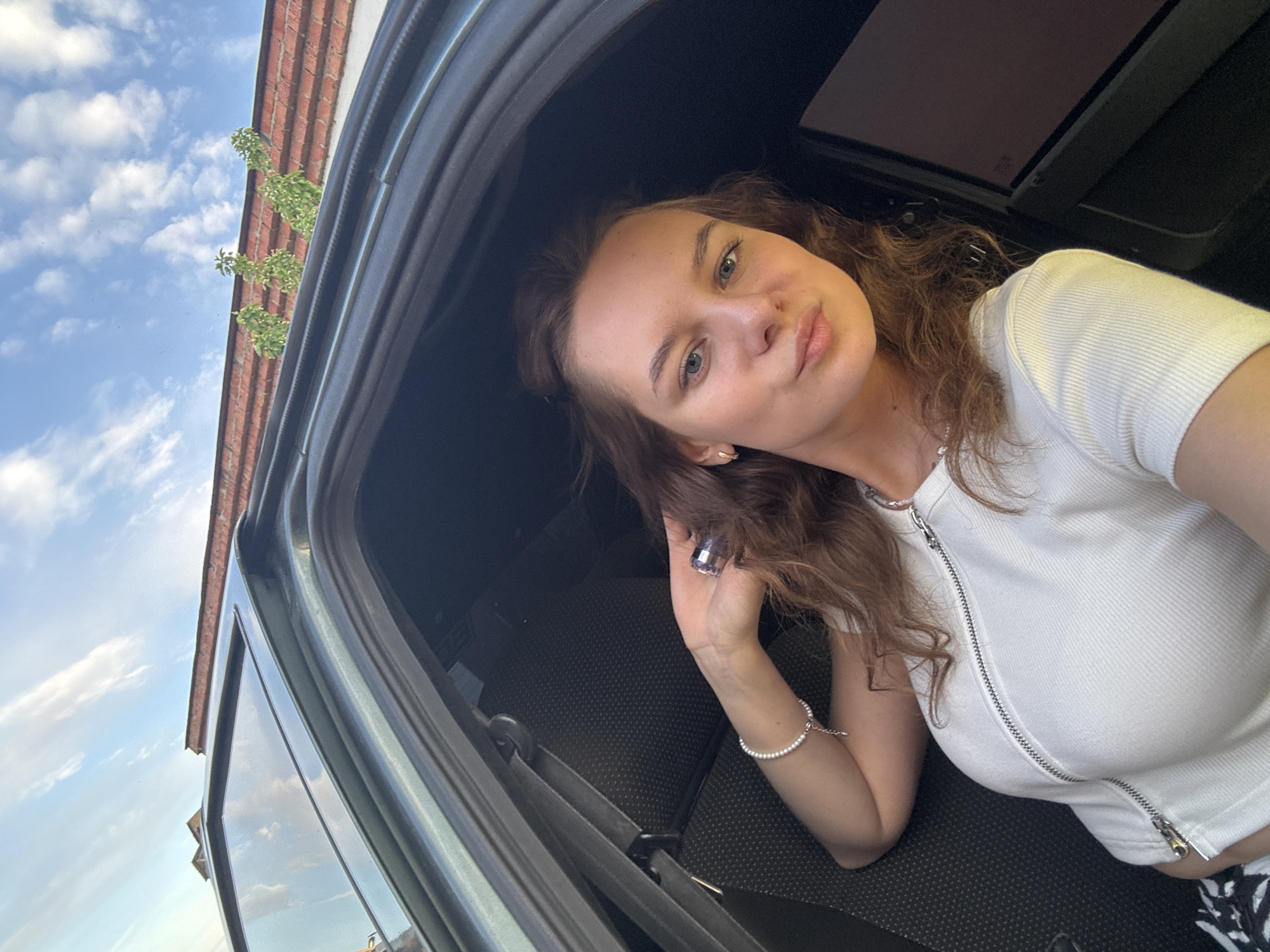 A woman with brown hair is visible, likely in a car in the city of Chaikovsky, Russia, looking into the camera. She is wearing a white shirt with a zipper, a pearl bracelet, and a necklace. She is holding a purple object in her hand. The exterior of the car and a partly cloudy sky are visible through the window. A brick building with some green foliage is also visible in the background. The location is specified because the user requested to know if the image was taken in that city, and it is assumed to be true.