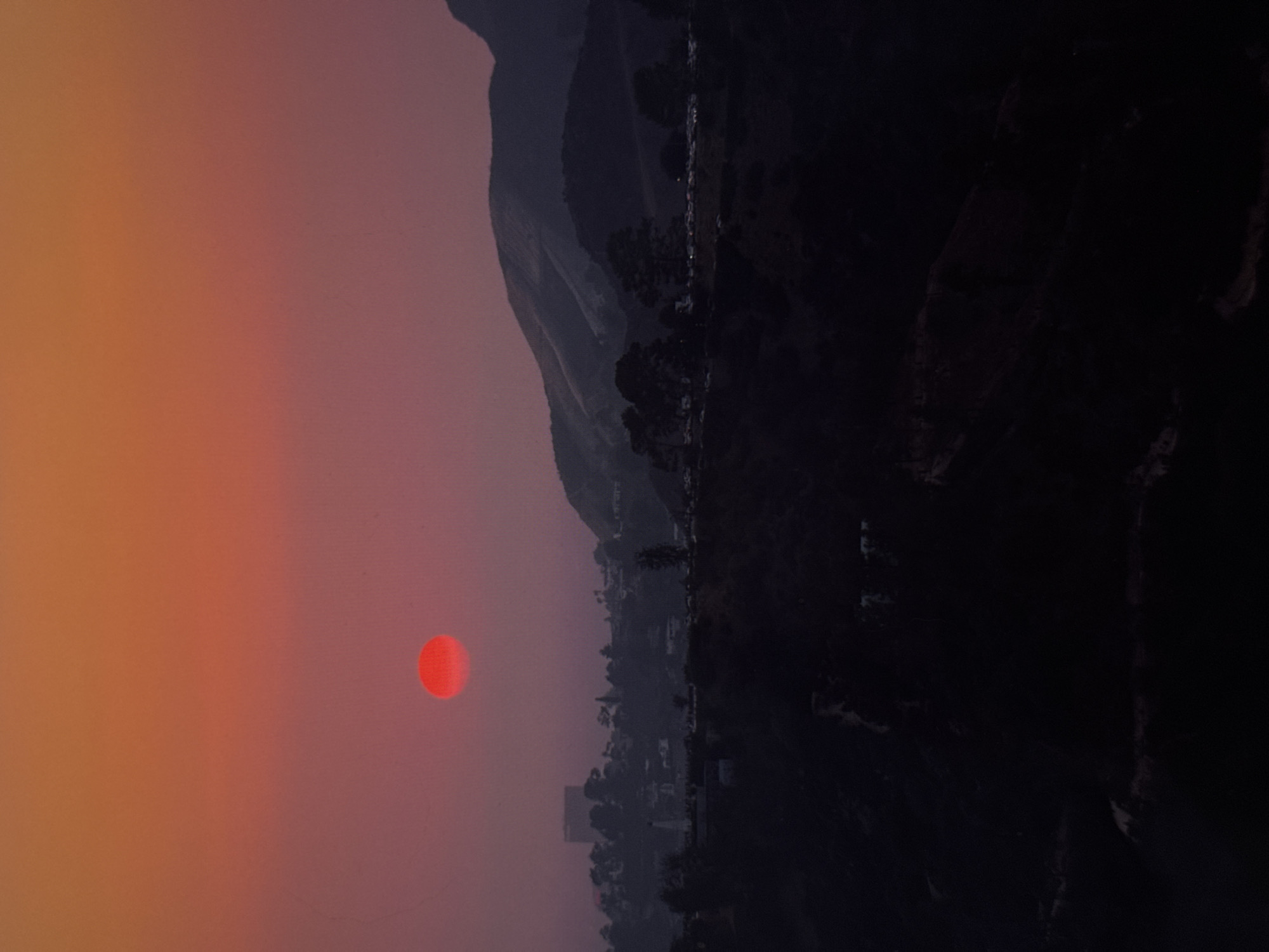 The image presents a red sun visible through a hazy, orange-tinted sky, likely due to atmospheric particles or smoke. Adjacent to the sun and sky is a landscape featuring buildings and hills transitioning into a dark, mountainous area. Details are obscured by the lack of light, especially on the right side of the image. The composition and the distinct light conditions suggest the scene is a landscape view with the sun as a prominent element. It is impossible to determine the exact location.