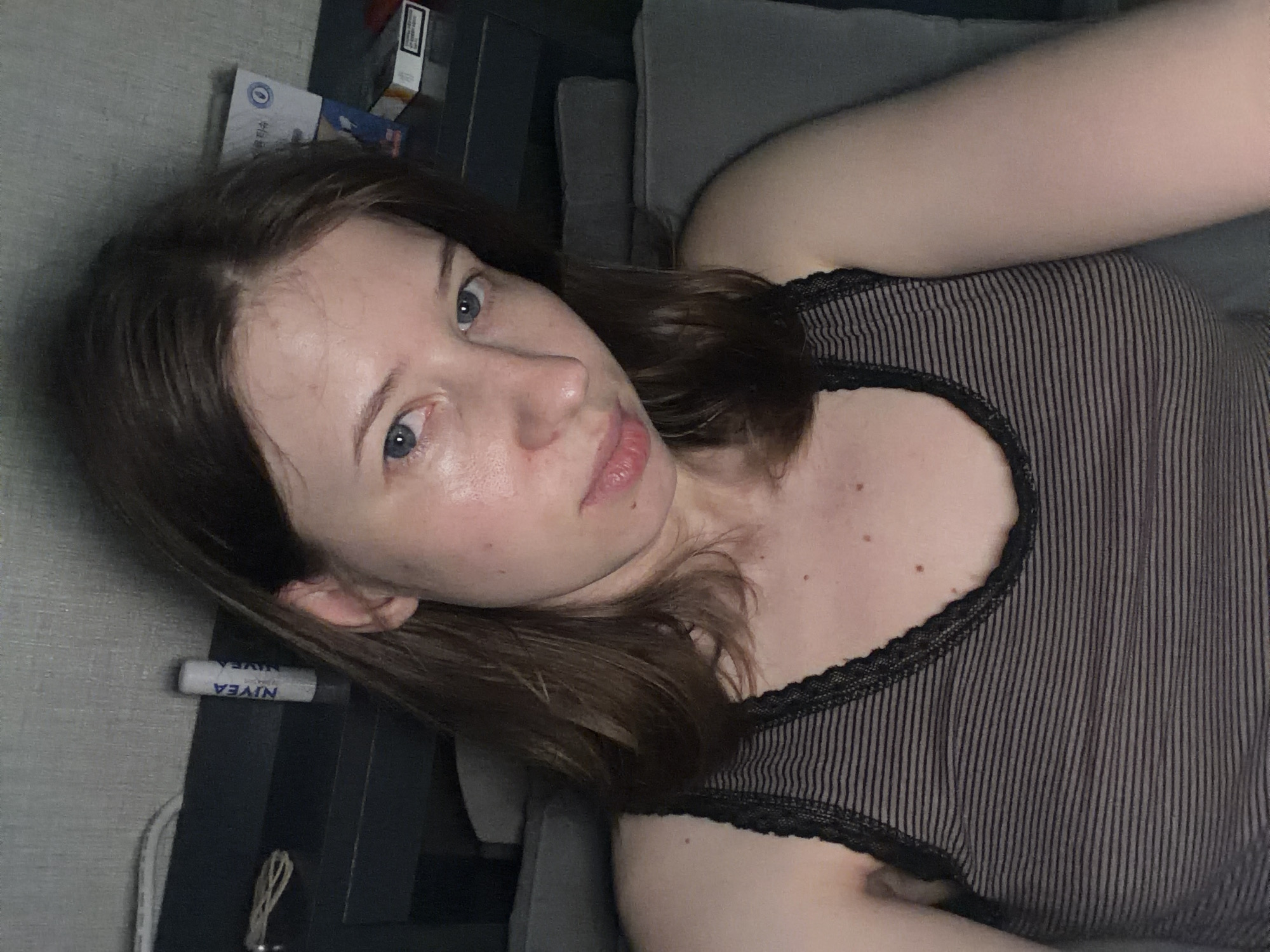 A young woman with light skin and brown hair is pictured in a close-up, selfie-style shot. She is wearing a black and grey striped tank top with lace trim. The background appears to be a bedroom, with a headboard and pillows visible. On the headboard, a Nivea lip balm and a pack of cigarettes are present. The lighting is dim, suggesting it may be nighttime or a low-light environment. There is no identifiable information that can place this image in Dongducheon-si, South Korea.
