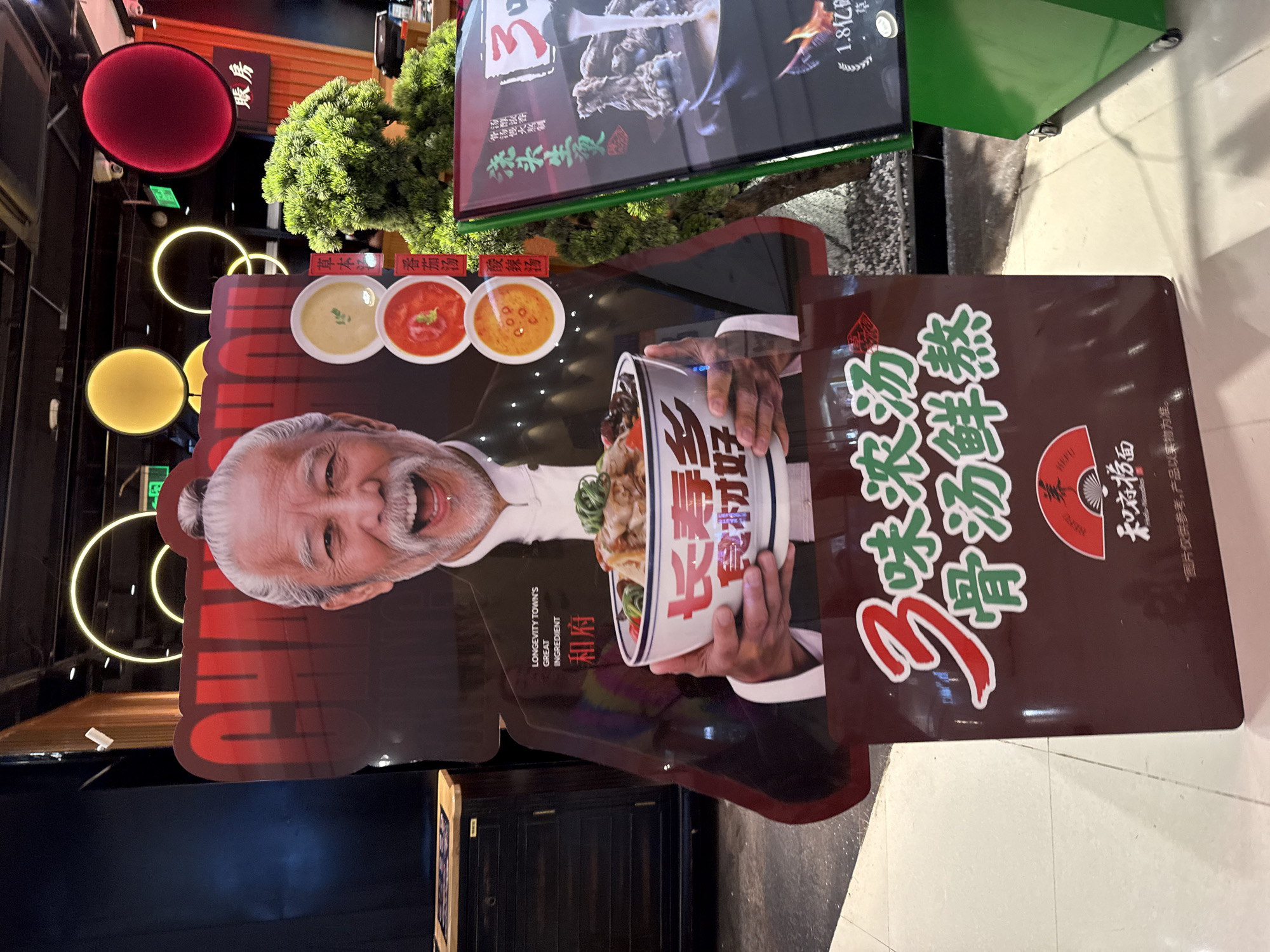 The image shows a life-sized cardboard cutout of an elderly Asian man smiling and holding a large bowl of noodles. The cutout is placed in front of a restaurant in Huizhou, China. Behind the cutout, there is a decorative plant and a sign for the restaurant. The sign features Chinese characters and images of food. The overall scene suggests a promotional display for a restaurant, likely a noodle shop, in a commercial area. The cutout's smiling expression and the appetizing food imagery are intended to attract customers.