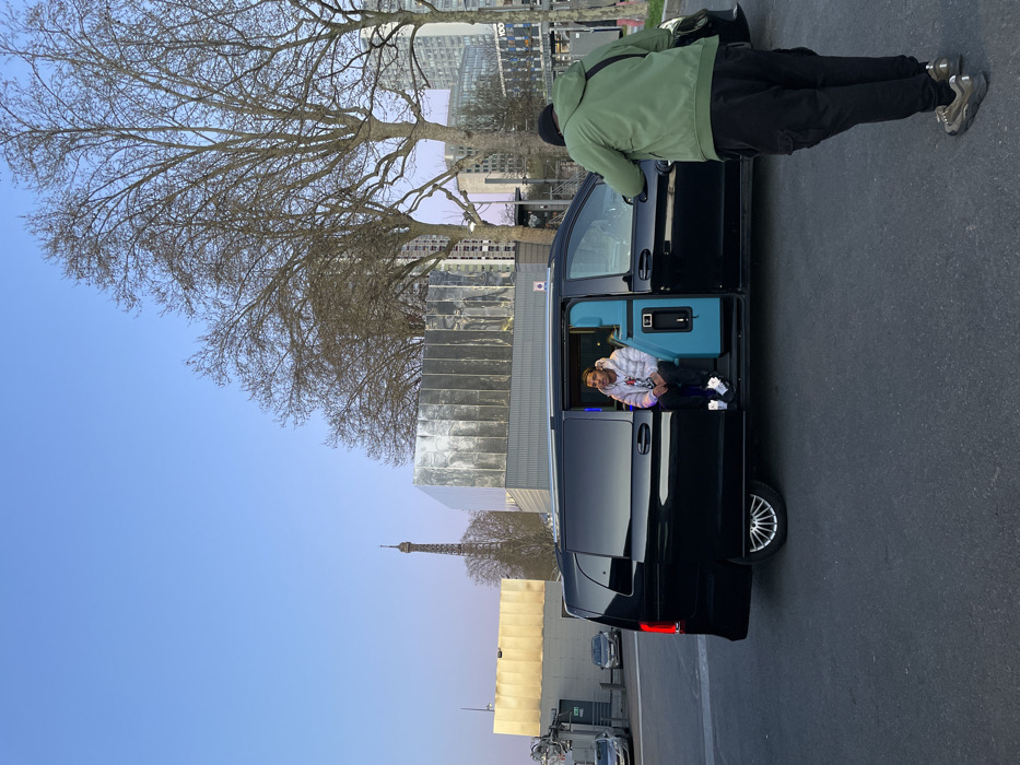An outdoor scene in Paris, France, captures a posed photograph in an urban setting. The Eiffel Tower is visible in the distant background on the left, silhouetted against a clear, light blue sky. In the midground, a black luxury van (likely a Mercedes-Benz V-Class) is parked on a dark paved surface. Its passenger-side sliding door is fully open, revealing a vibrant turquoise interior lining. A male individual is seated inside the van, facing outward, wearing a white patterned hooded jacket, dark pants, and white sneakers. To the right of the van, another male individual stands with his back to the viewer, leaning forward, holding a device (likely a smartphone or camera) aimed at the seated person. He is dressed in a dark green jacket, dark cargo pants, and light brown sneakers. The background features several bare deciduous trees and various modern buildings with reflective metallic and golden facades, typical of areas like the Quai Branly or near the Palais de Tokyo.