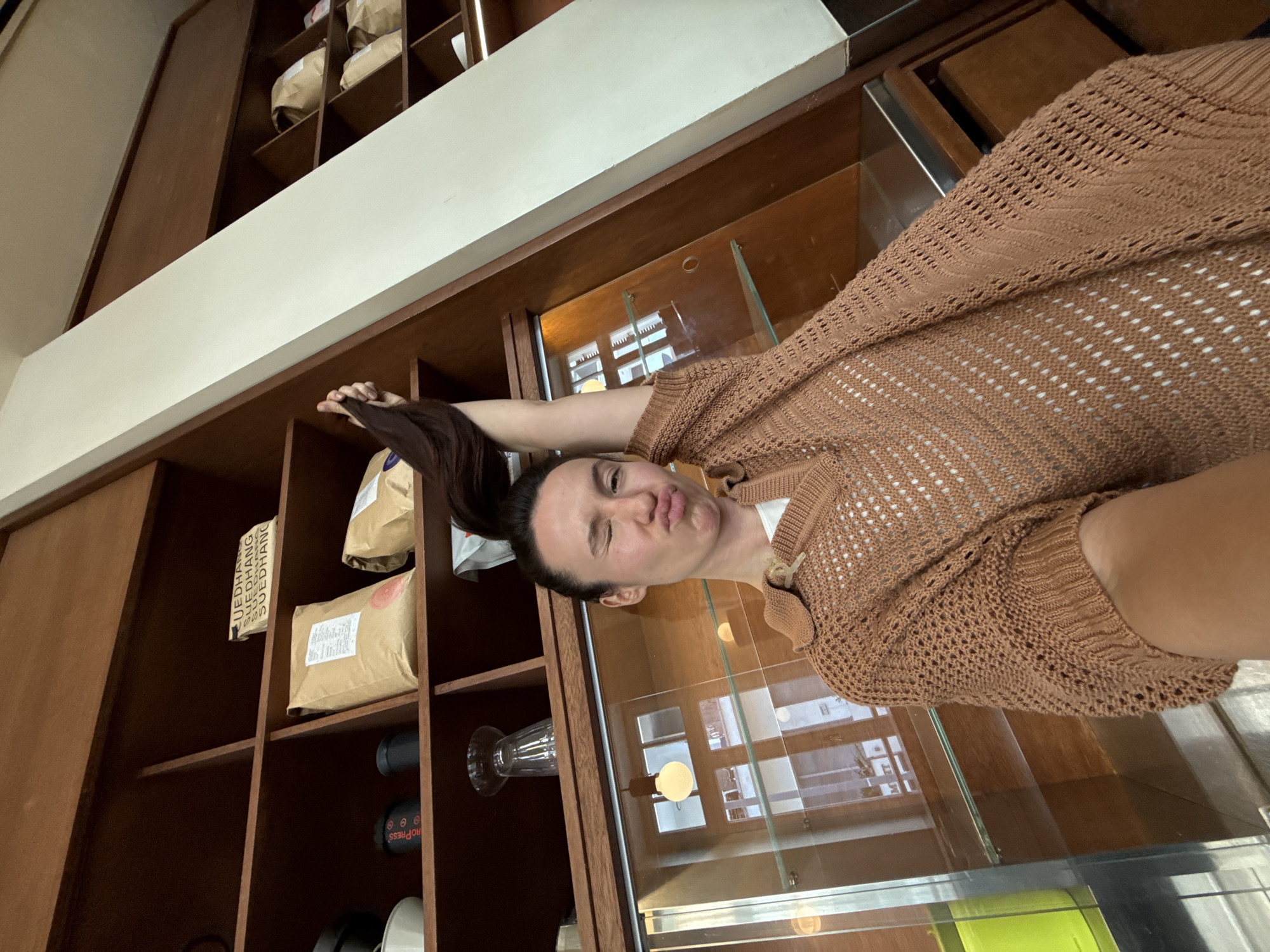 A woman is taking a selfie in a cafe, holding her ponytail up in the air. The woman has dark hair pulled back in a ponytail and is wearing a brown, knit sweater over a white shirt. She is making a kissing face toward the camera.
The background includes a wooden display cabinet with shelves containing coffee bags and accessories. A glass display case is adjacent to the shelves, reflecting windows and lights.
Based on the style of the cafe and available information, the location may be in Tel Aviv, Israel.