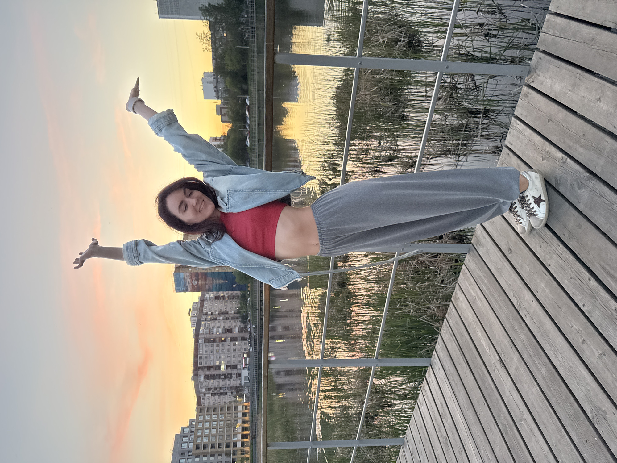 The image captures a young woman standing on a wooden pier, likely in the city of Tyumen, Russia, given the architectural style of the buildings visible in the background. The pier is constructed of light-colored wooden planks laid out in parallel. The woman is striking a pose with her arms outstretched, appearing to be embracing the surroundings. She is wearing a red top, loose grey pants, a light blue denim jacket, and white sneakers adorned with stars. Behind her, a body of water reflects the surrounding buildings and the sky, which is bathed in the warm hues of a setting or rising sun. A railing, made of grey metal, runs alongside the pier, separating it from the water. In the background, urban buildings are visible against the sky.
