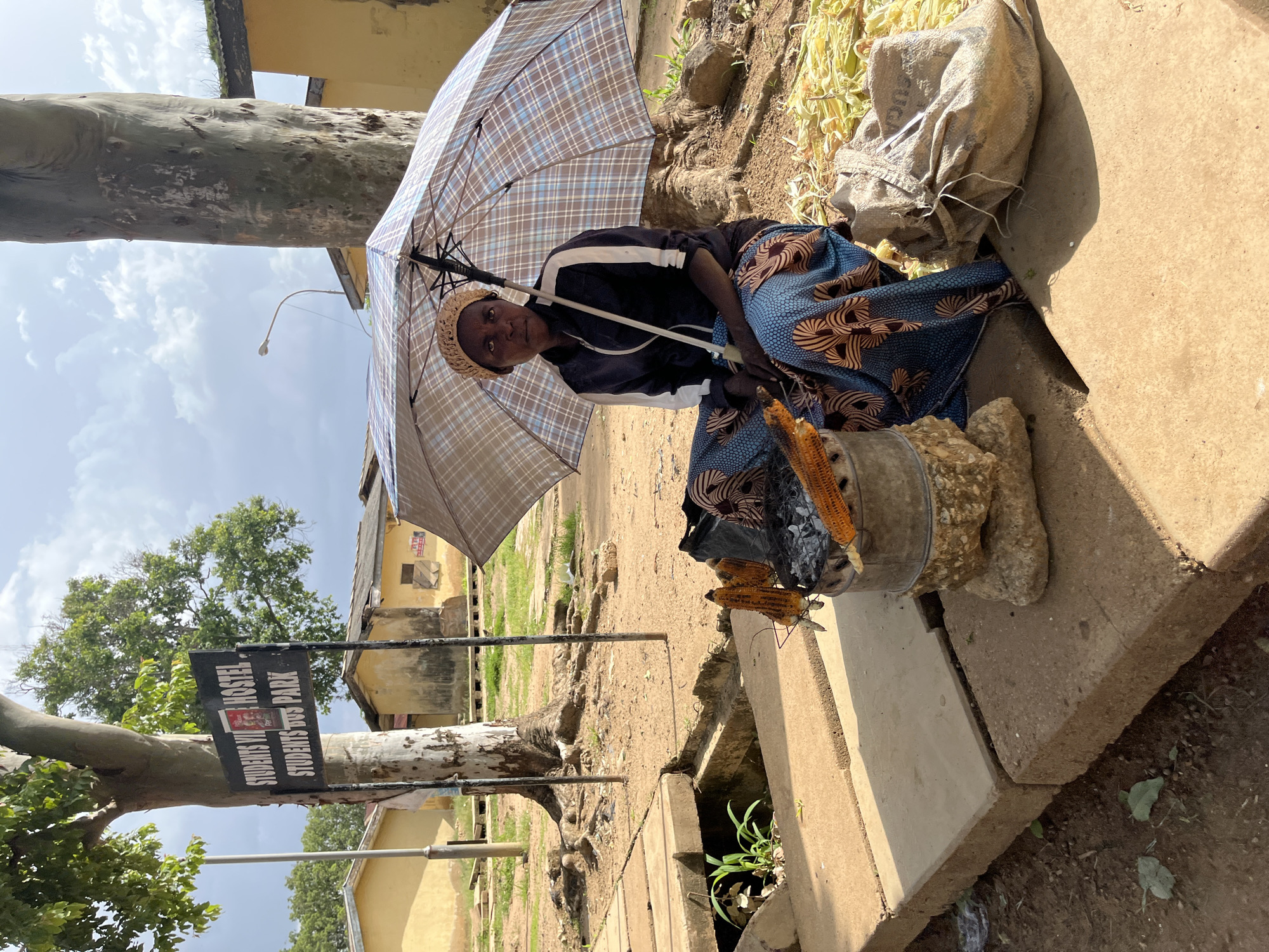 A woman sits under a plaid umbrella, holding a piece of corn. She is dressed in a blue and brown patterned skirt and a dark jacket with white stripes. Beside her is a metal bucket filled with corn cobs. A large sack, likely containing more corn, is behind her, and to her left, a sign reads "STUDENTS' VI HOSTEL STUDENTS' BUS PARK". The scene appears to be outdoors, with a dirt path and some greenery. A large tree trunk is visible on the left side of the image. The lighting suggests it is daytime. The woman's expression is neutral.