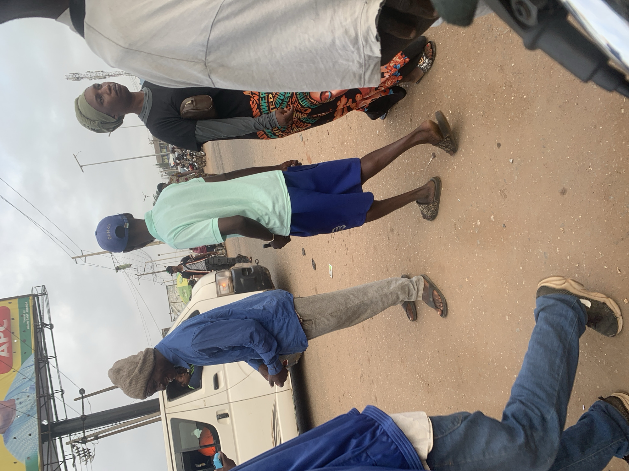 In Shagamu, Nigeria, a group of people are visible on a dirt road under an overcast sky. A white van is parked on the left, with its driver's side window open. A man wearing a blue shirt and grey pants, with a knitted beanie, stands near the van, looking towards the left and smiling. To his right, a person in a light blue t-shirt and blue shorts is visible, looking forward. Further to the right, a woman in a black t-shirt and a green headscarf is seen with a small brown bag. Behind her, another person in a white shirt is partially visible. In the background, a billboard featuring a man in a blue suit with the "APC" logo is mounted on poles. Traffic lights and electrical poles are also present in the background, suggesting an urban environment. The ground is a mixture of sand and dirt, with scattered debris.