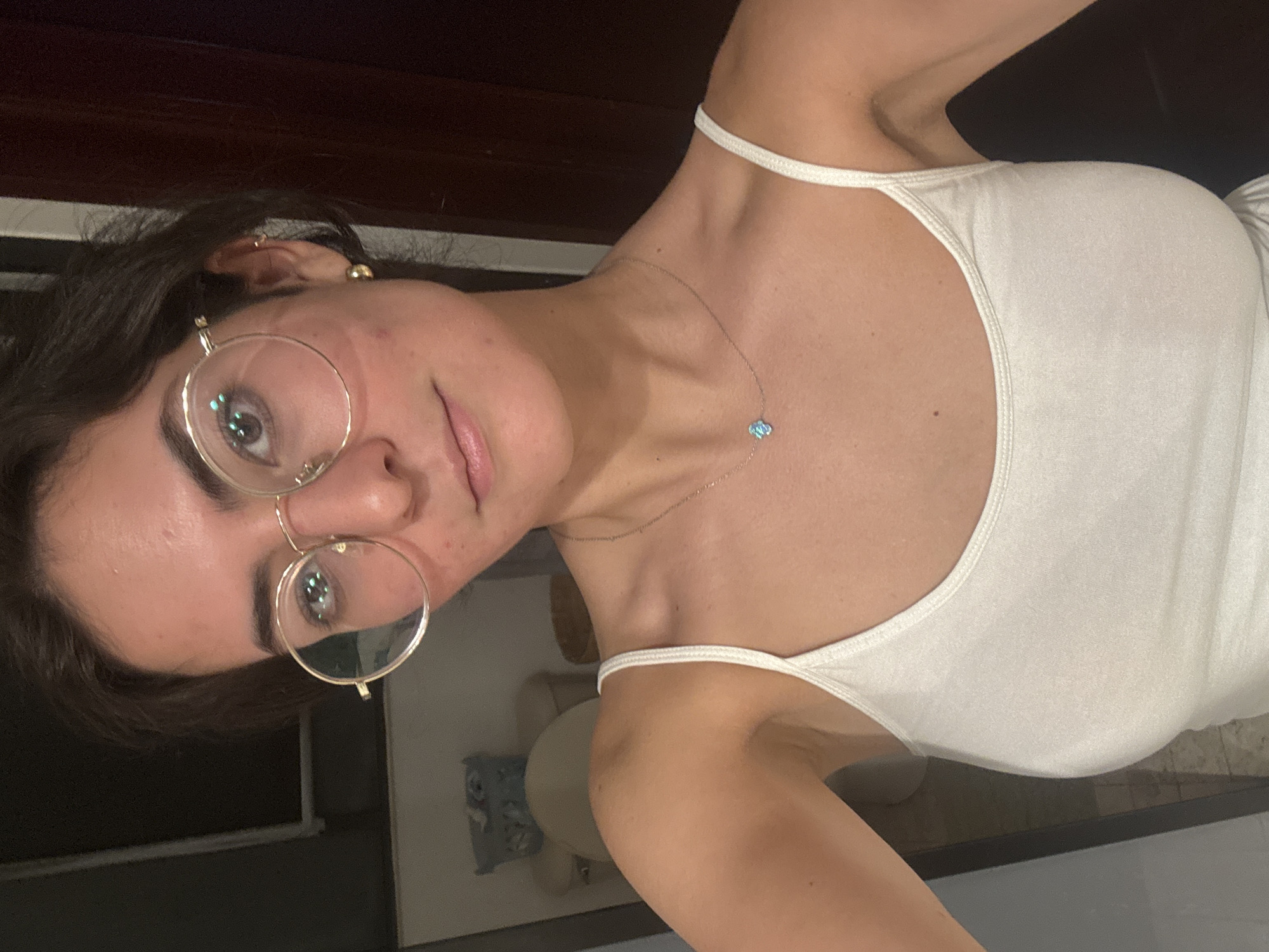 A person is posing indoors. The person has brown hair, wears round glasses, and has a necklace with a small blue pendant. The person wears a white tank top. The location appears to be inside a residential dwelling. The city of Puebla, Mexico, cannot be determined from this image.