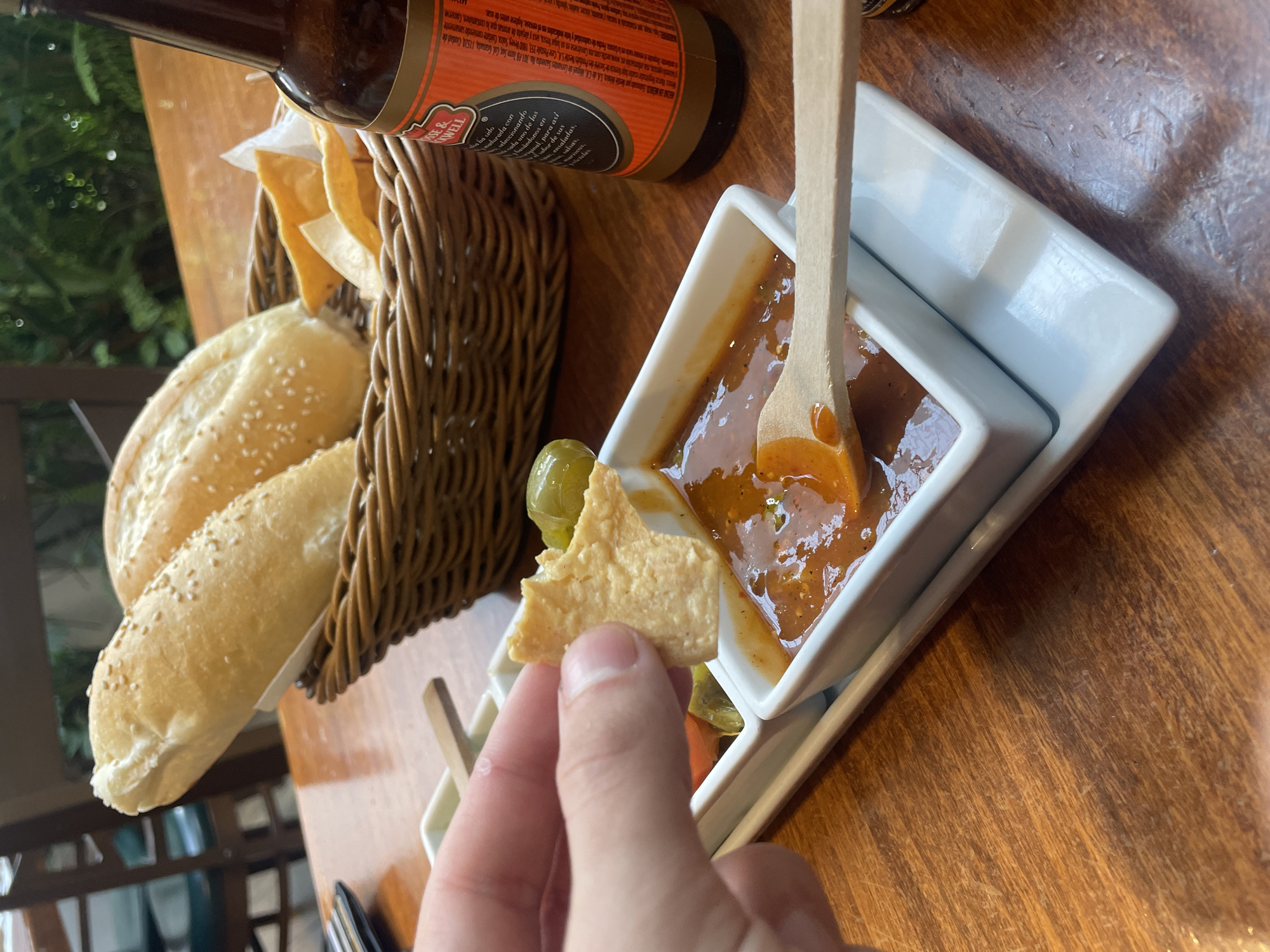A close-up shot captures a hand dipping a tortilla chip into a white rectangular dish filled with a vibrant orange salsa, set against a wooden table. The chip, already partially submerged, is angled to scoop up the salsa. Adjacent to this dish, another smaller white dish holds a few green olives. In the background, a woven basket overflows with two long, lightly browned bread rolls sprinkled with sesame seeds, and a crumpled white napkin. To the right, the neck and label of a dark glass beer bottle are visible. The environment suggests an indoor or semi-outdoor dining setting with diffused natural light illuminating the scene.